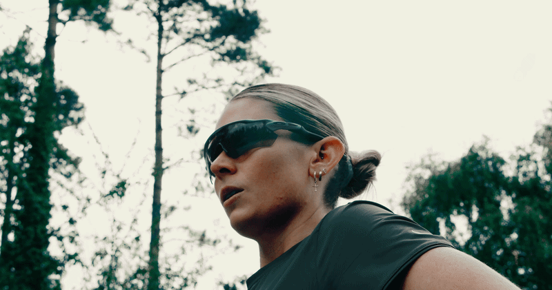 A woman with a sleek ponytail wearing dark sunglasses and earrings, outdoors among tall trees on a cloudy day.