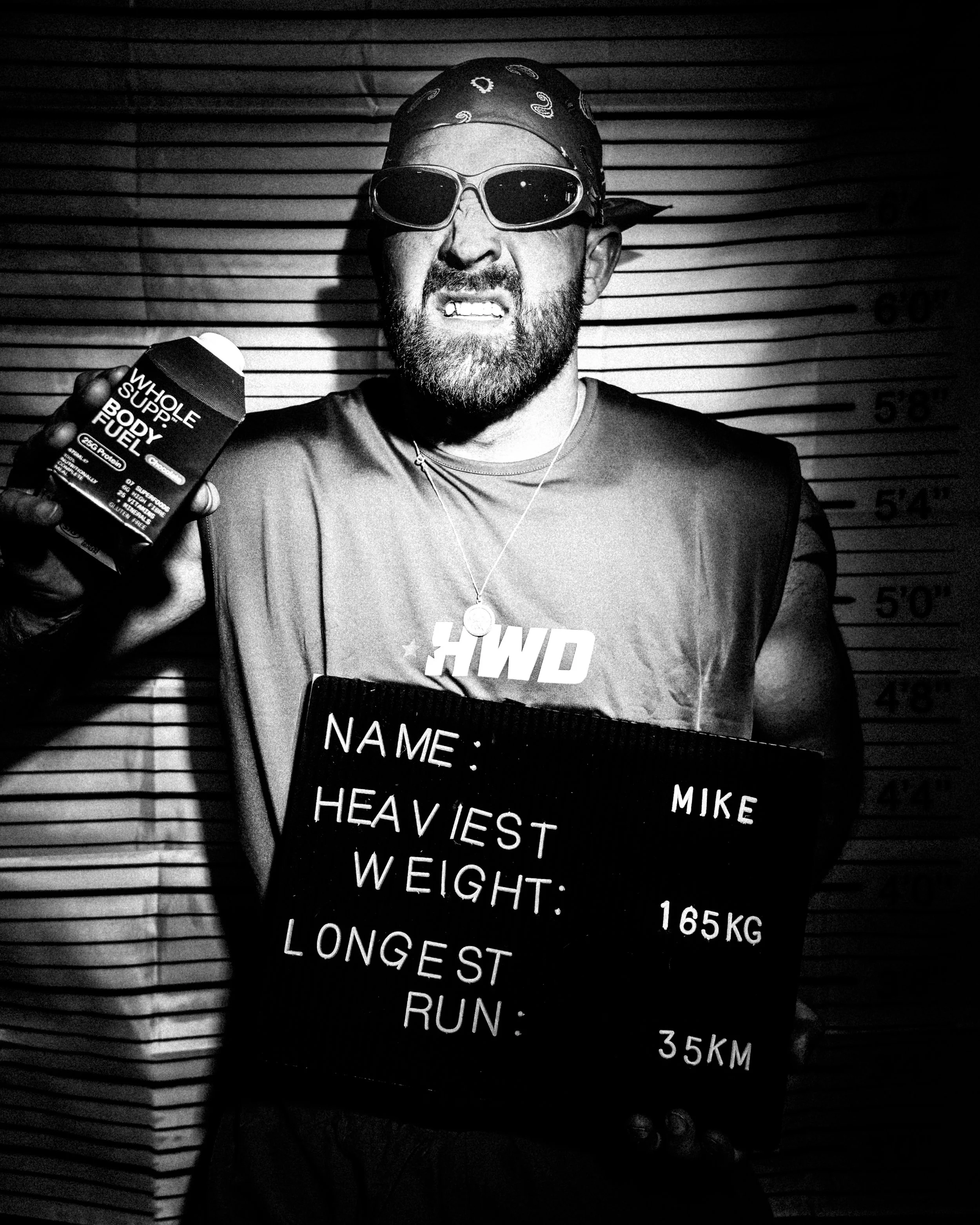Black and white photo of a man wearing sunglasses, a bandana, and a t-shirt, holding a bottle of supplement labeled 'Whole Supp. Body Fuel'. He is standing in front of a height measurement wall, with a sign in front of him displaying his name and weight.