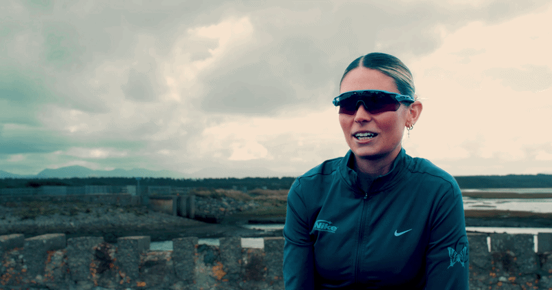 A woman wearing sunglasses and a blue Nike jacket outdoors, with cloudy skies and water in the background.