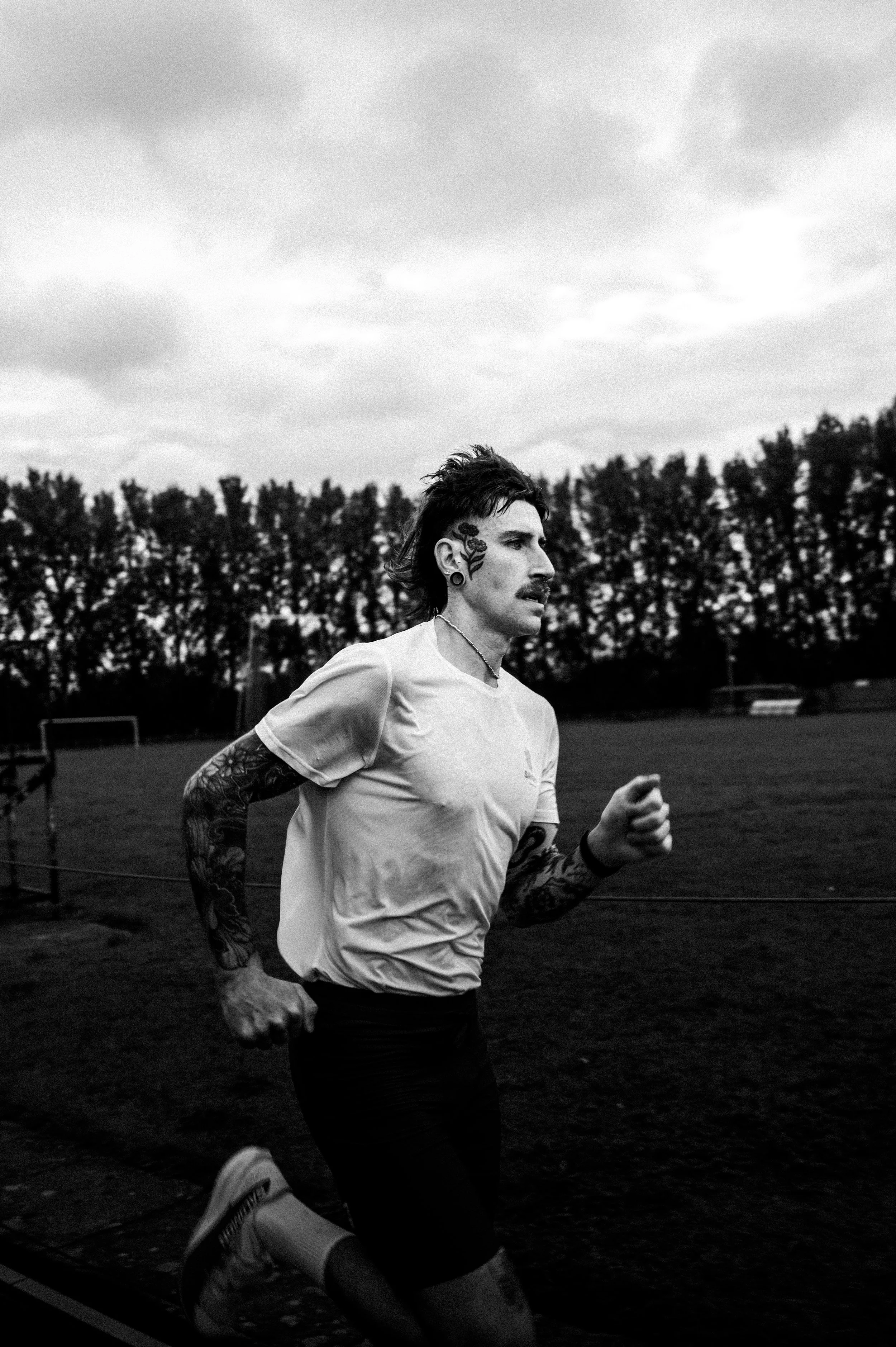 A man with tattoos on his arms and face, wearing a white shirt and running outdoors on a field or track with trees in the background and cloudy sky.