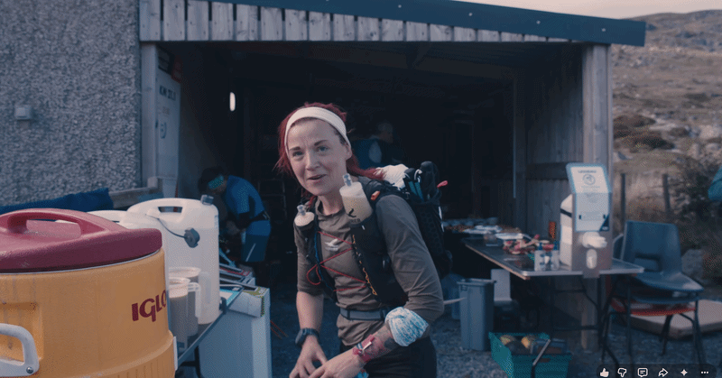 sanna duthie with a salomon running vest and headband standing at an aid station during the uts100 race near a small shelter with other people and supplies in the background.
