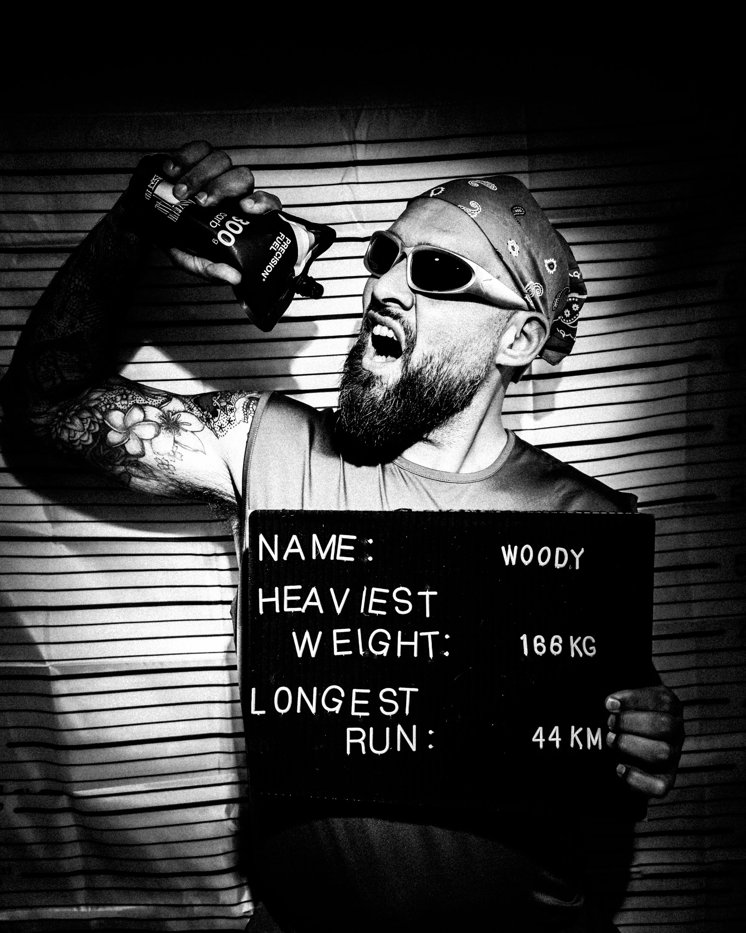 Black and white photo of a man with a beard, wearing sunglasses, a bandana, and a sleeveless shirt, taking a selfie with a paddleboard number board showing the highest weight and longest run in a lineup of other sheet boards.