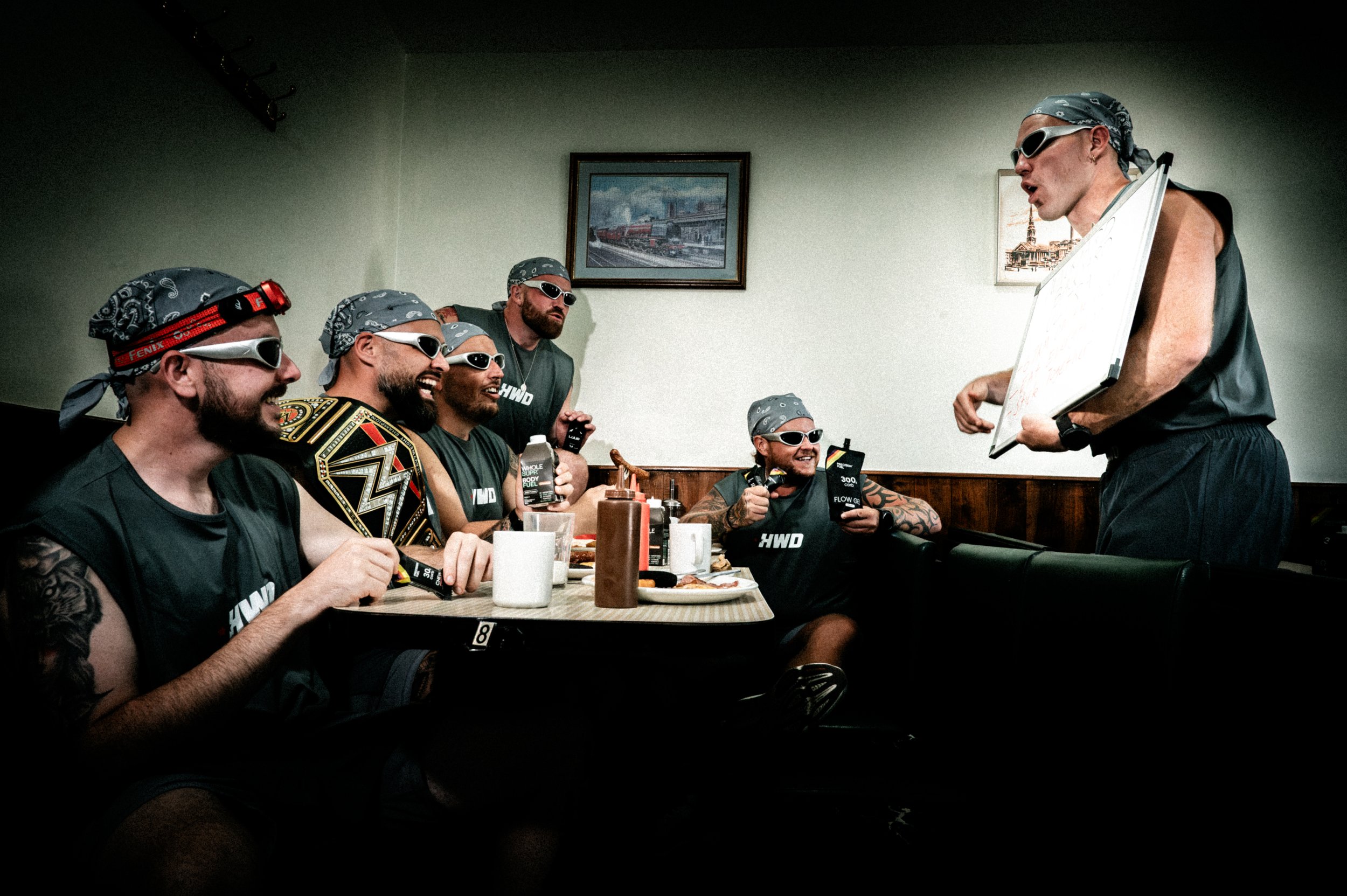 A group of five men dressed in black t-shirts and bandanas, wearing sunglasses, sitting at a dining table in a restaurant, showing excitement and laughter while looking at a man standing with a whiteboard, who appears to be talking to them. The table is set with food, drinks, and condiments, and there are framed pictures of trains and landmarks on the wall behind them.