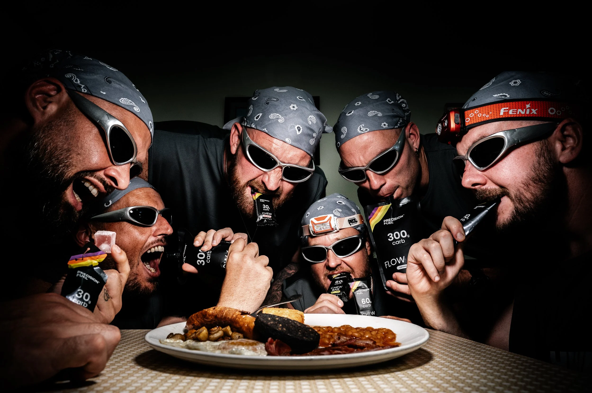 Group of men wearing sunglasses and bandanas, celebratorily eating food at a table with a plate of assorted foods, including meats and vegetables.