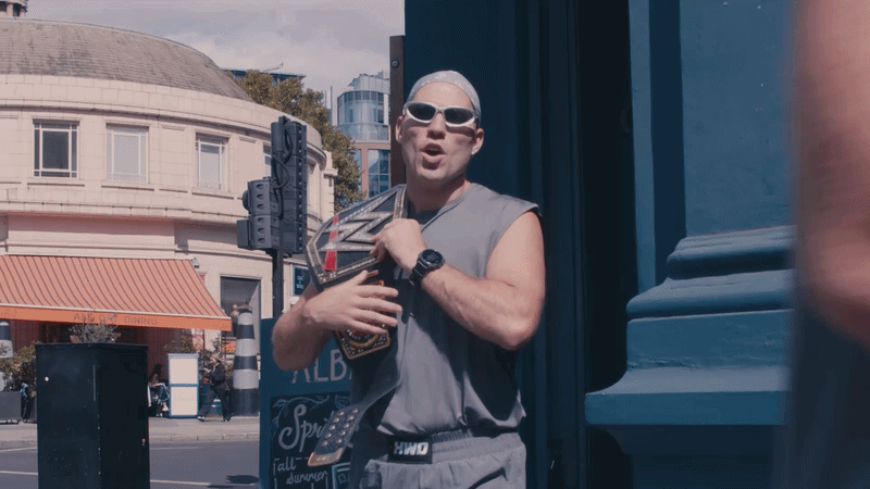 jordan stanton wearing sunglasses and a gray sleeveless shirt holding a WWE championship belt across his chest on a city street.