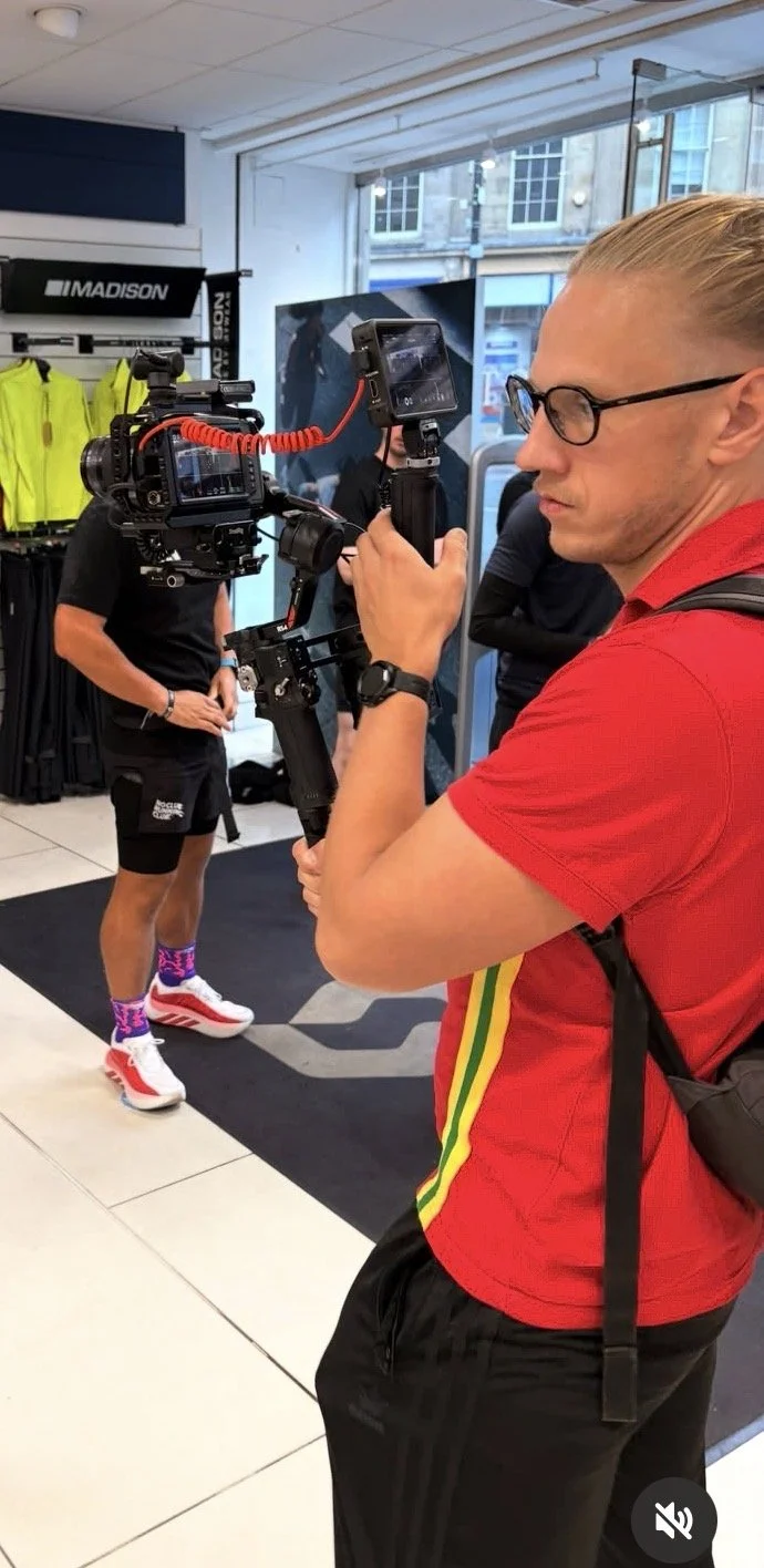 A man wearing glasses in a red shirt with yellow and green stripes on the sleeve is filming with a professional camera inside a retail store. In the background, a person in athletic gear and colorful socks is visible, along with bright yellow jackets on display.