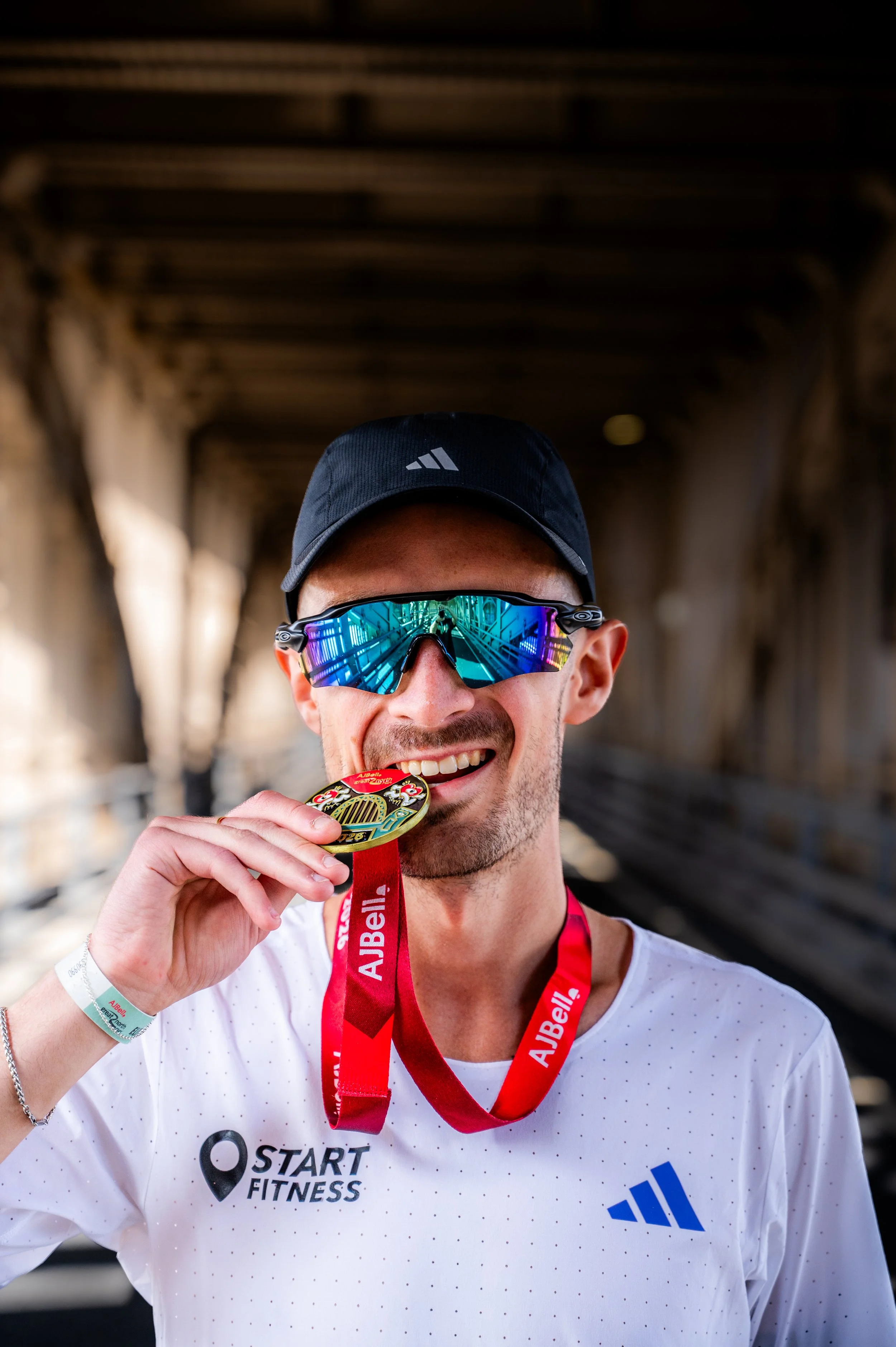 cole gibbens wearing an adidas black cap, reflective sunglasses, and a white start fitness and adidas shirt, smiles while holding up a gold medal with a red ribbon at an outdoor event under a bridge.