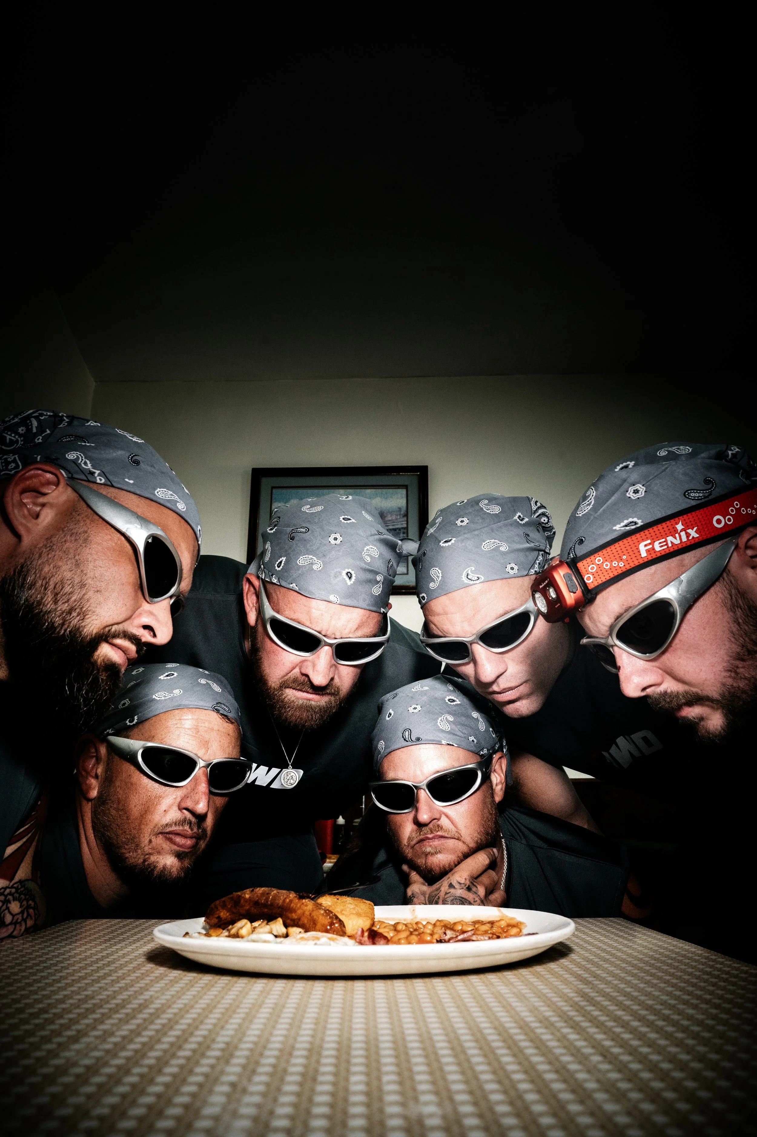 Group of men wearing sunglasses and bandanas gathered around a table with a plate of food, in a room with a picture hanging on the wall.