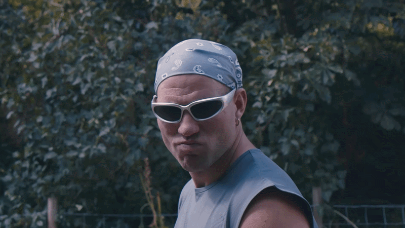 jordan stanton wearing a gray bandana and white sunglasses looks sternly at the camera with greenery in the background.