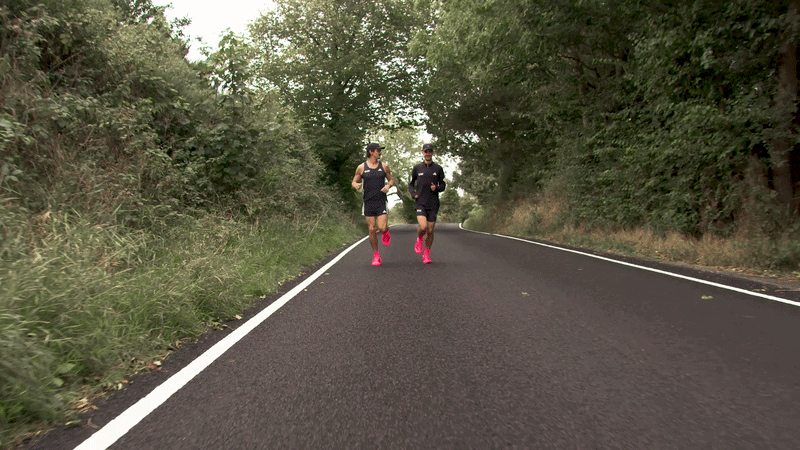 Two runners, a man and a woman, jogging on a winding road surrounded by trees and greenery.