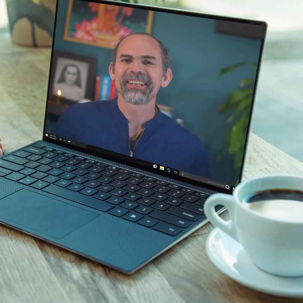Pantalla de computadora portátil que muestra a un hombre con barba y cabello corto en un fondo colorido, con una taza de café a la derecha en una mesa de madera.
