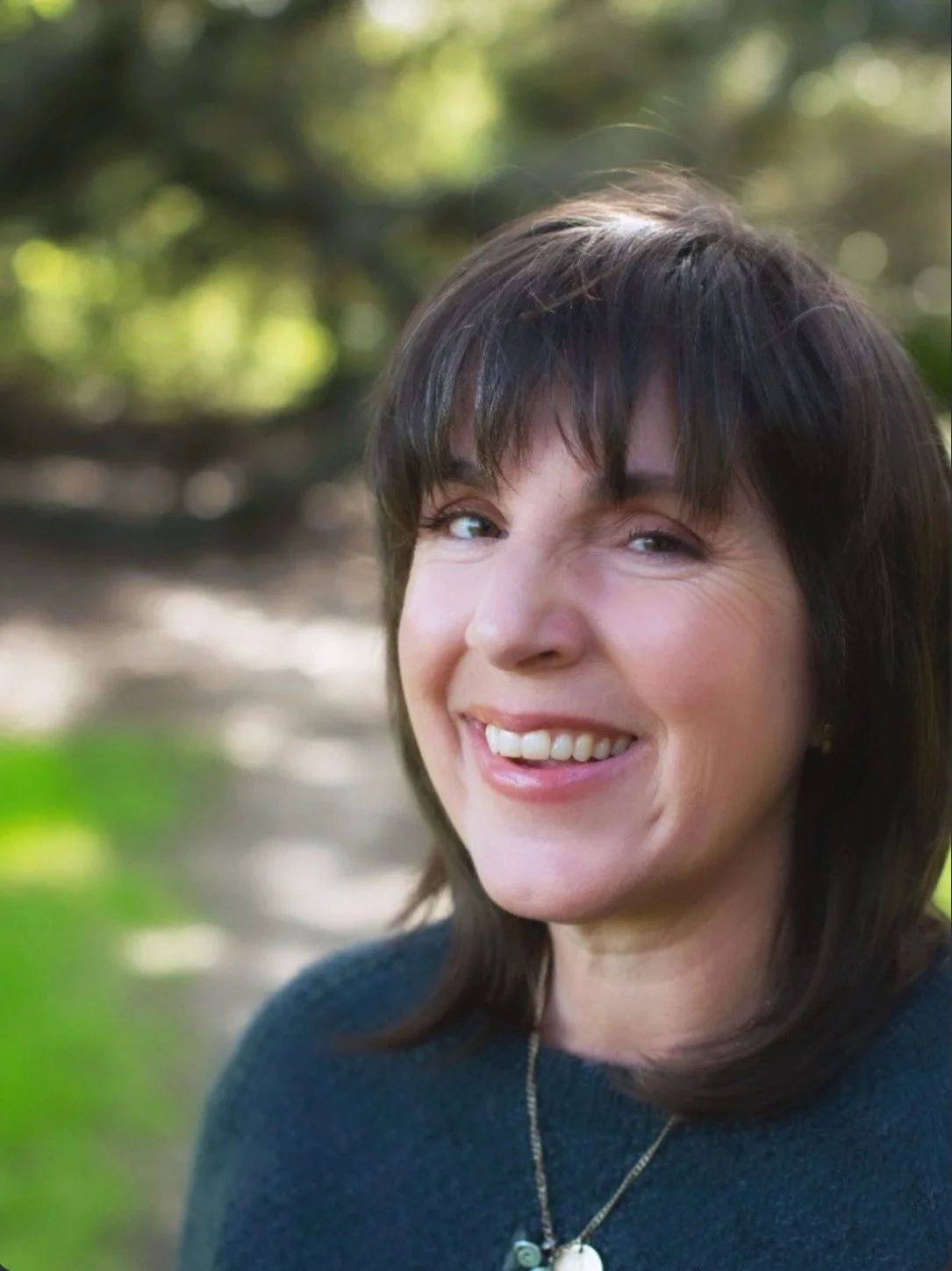 Woman (Alexis Scarborough) with long brown hair smiling outdoors wearing green sweater and pendant necklace