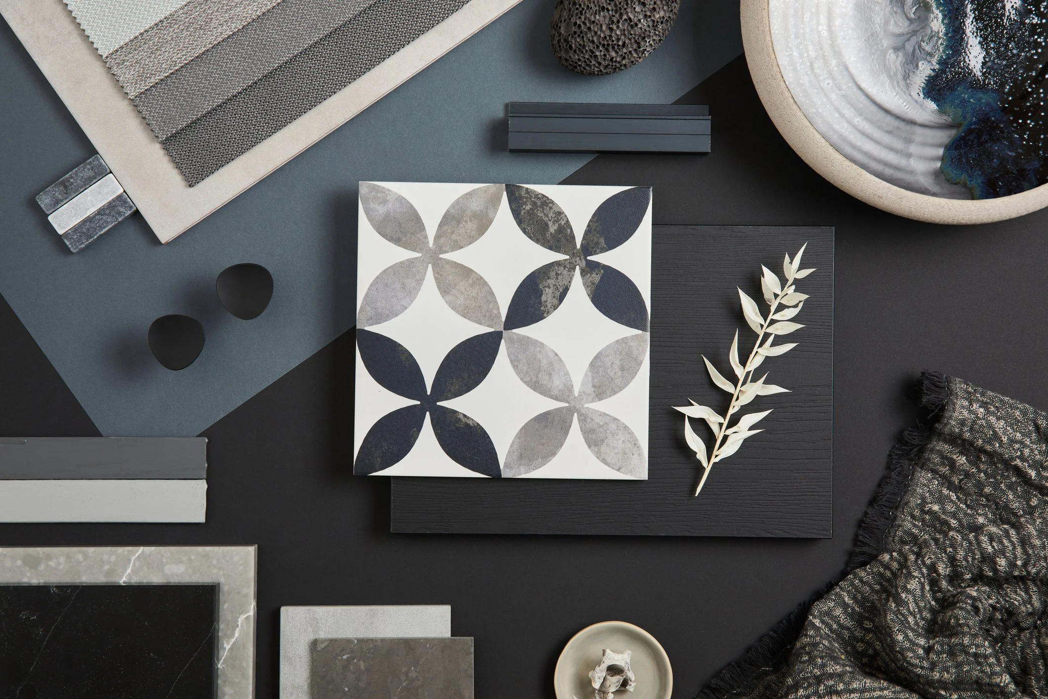 Flat lay of design materials including swatches, tiles, textured stone, ceramics, and fabric samples on dark surface.