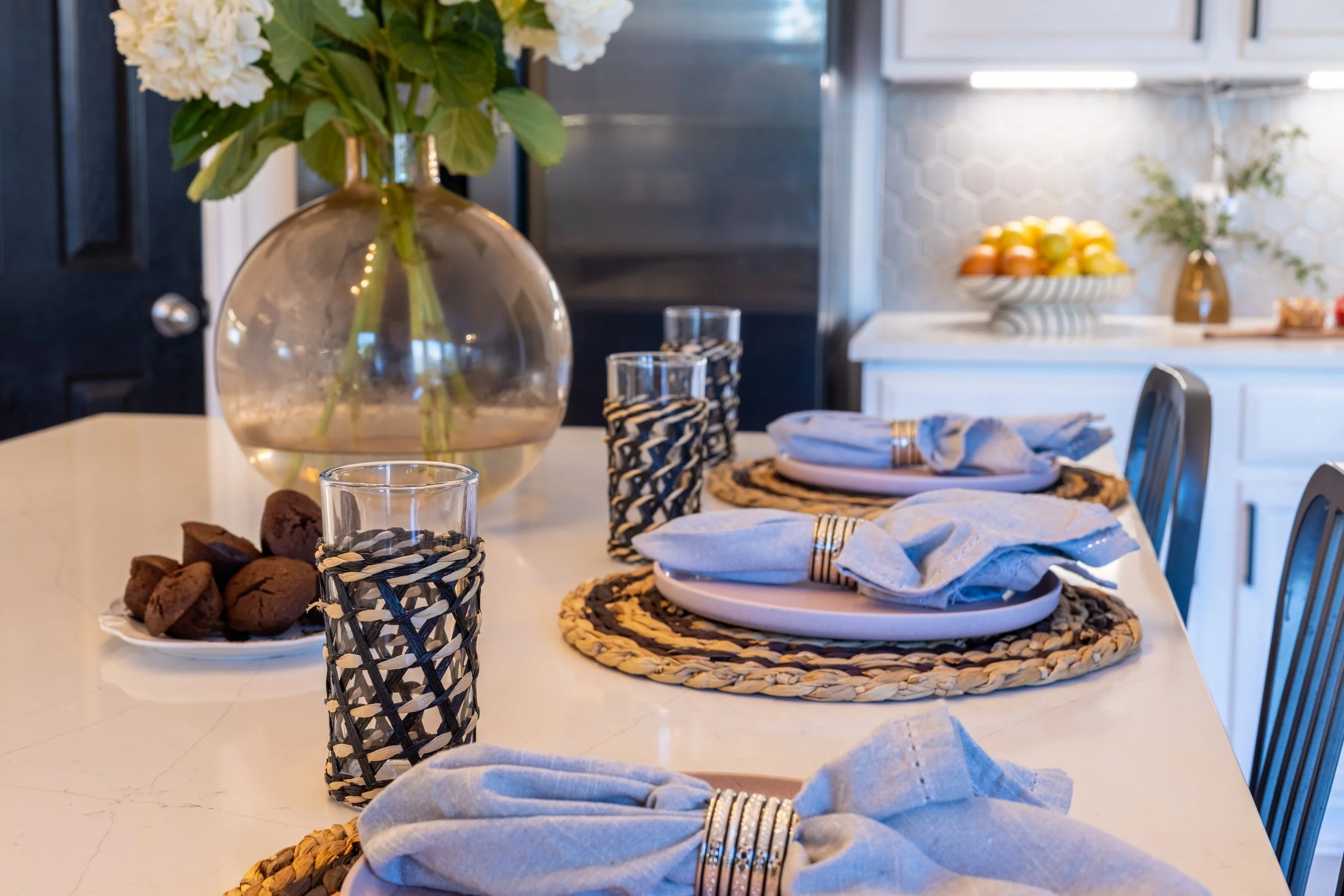 Dining table with pink plates, gold napkin rings, woven placemats, and floral centerpiece in a bright modern kitchen.