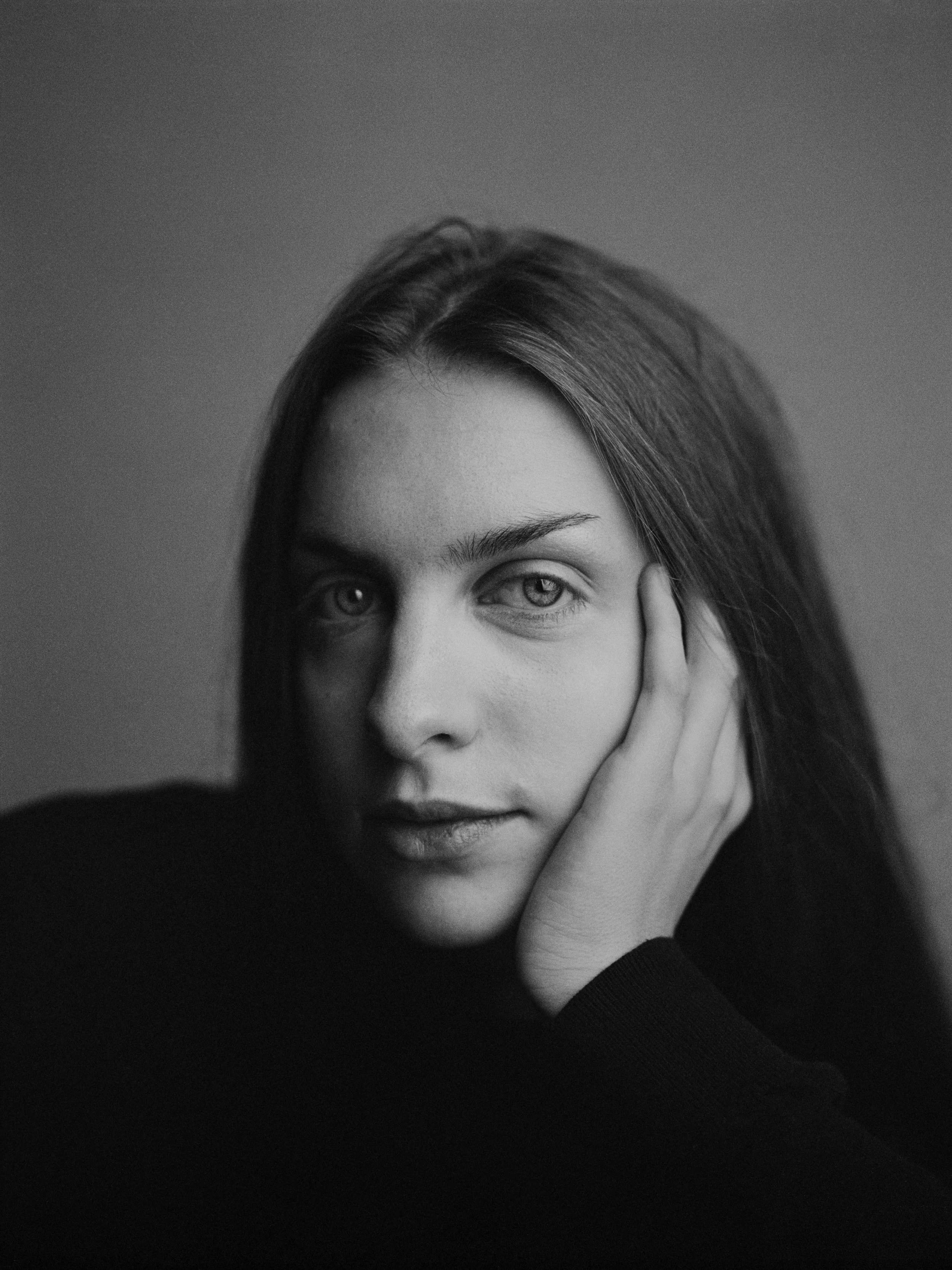 Schwarz-weiß Porträt einer jungen Frau, die ihre rechte Hand an ihr Gesicht legt und direkt in die Kamera schaut.