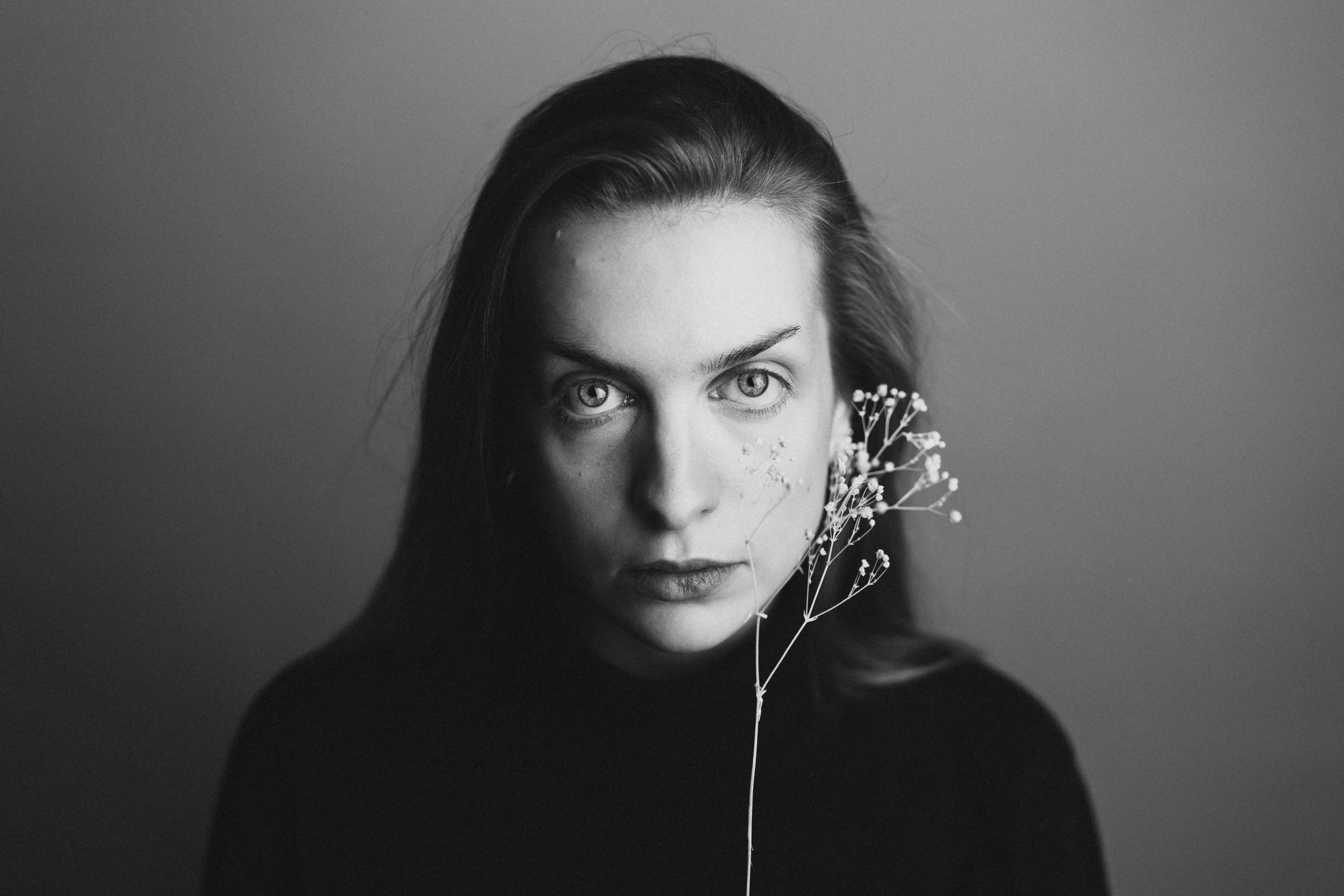 Schwarz-weiß Porträt einer jungen Frau mit dunklem Pullover, blickt direkt in die Kamera, hält eine kleine Blumenpflanze vor ihrem Gesicht.