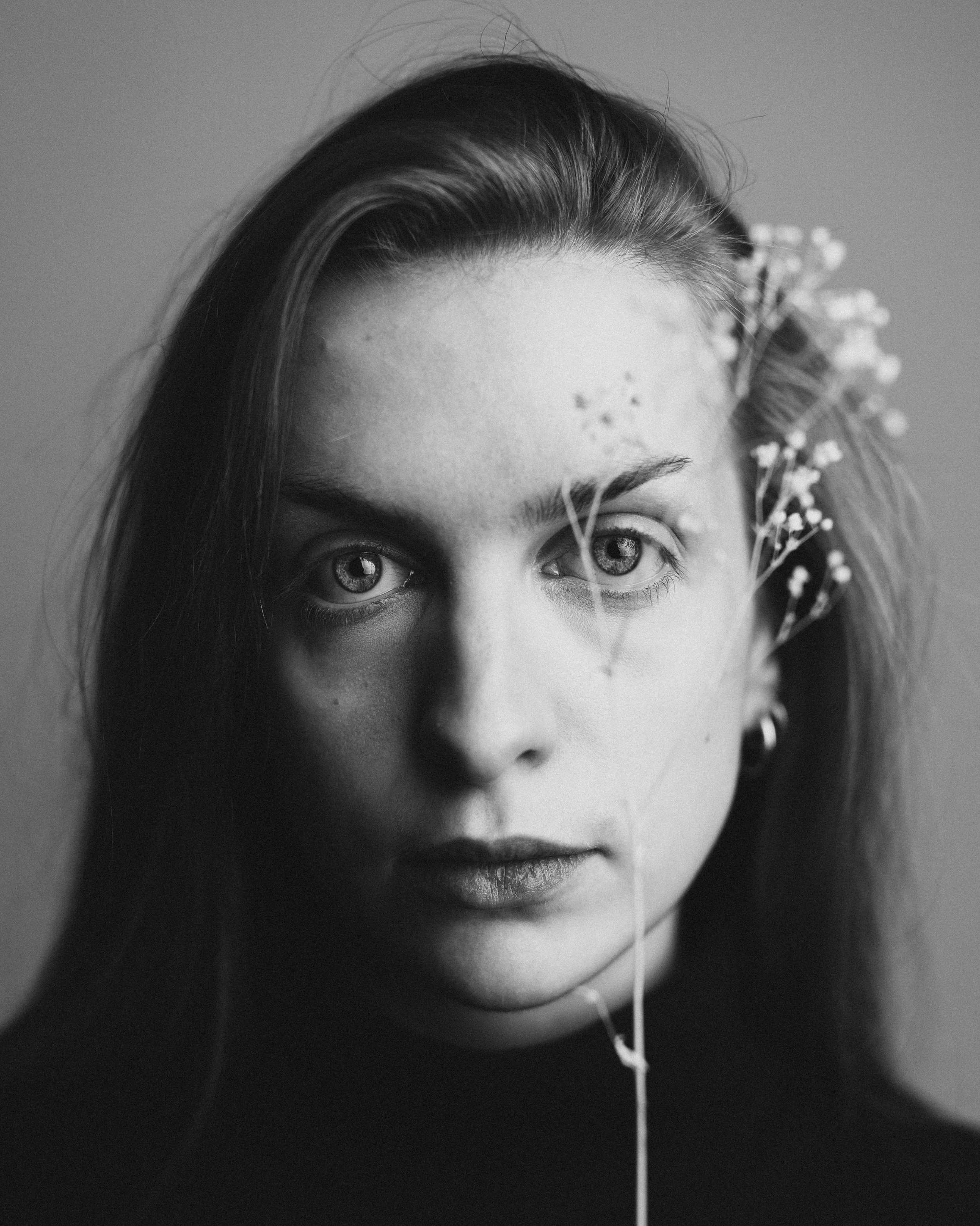 Schwarz-weiß Porträt einer jungen Frau mit langen Haaren, die direkt in die Kamera schaut, mit kleinen Blumen vor dem Gesicht.