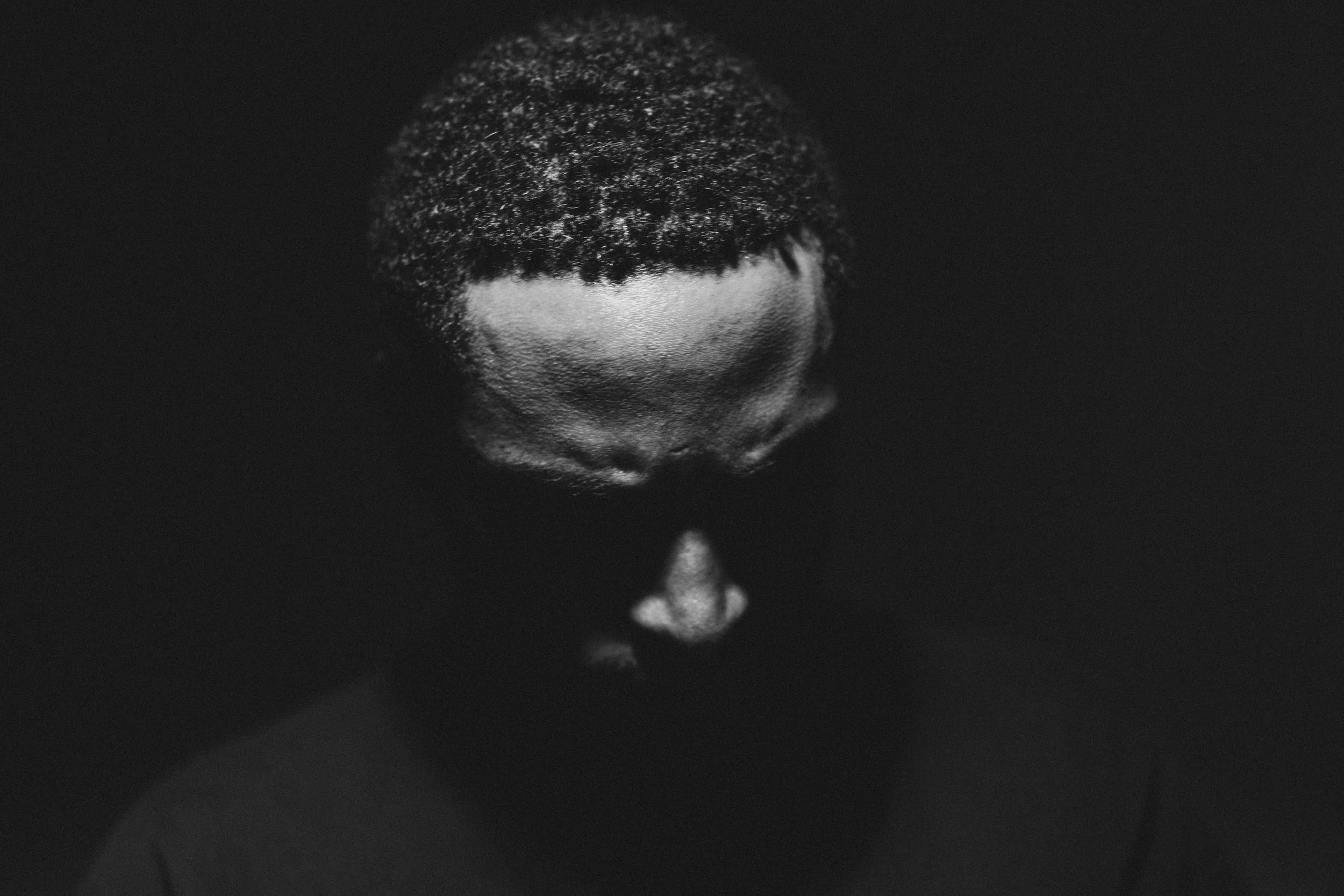 Ein schwarz-weiß Bild eines Mannes mit Kurzhaarschnitt, dessen Kopf nach unten geneigt ist, gegen einen dunklen Hintergrund