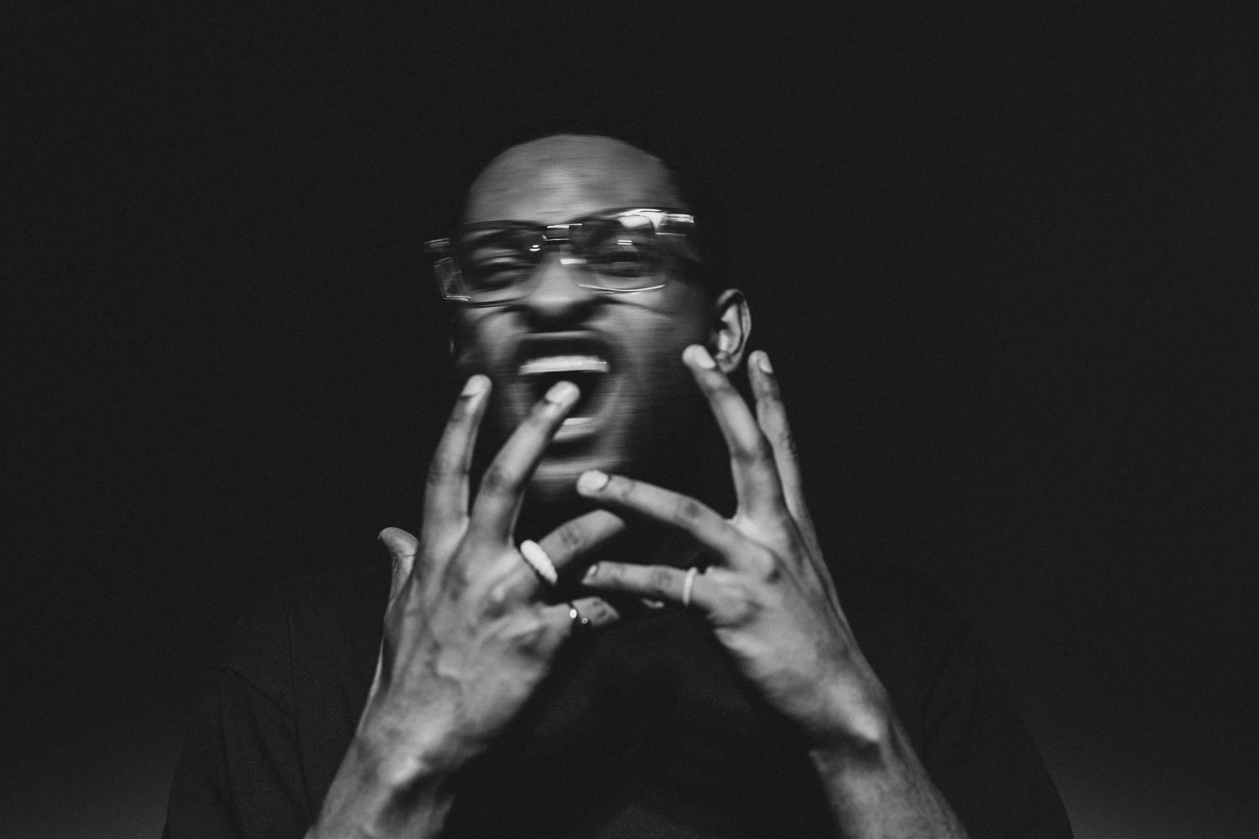 Schwarz-weiß Fotografie eines schwarzen Mannes mit kurzen Haaren und Brille, der seine Hände vor den Mund hält, während er schreit.