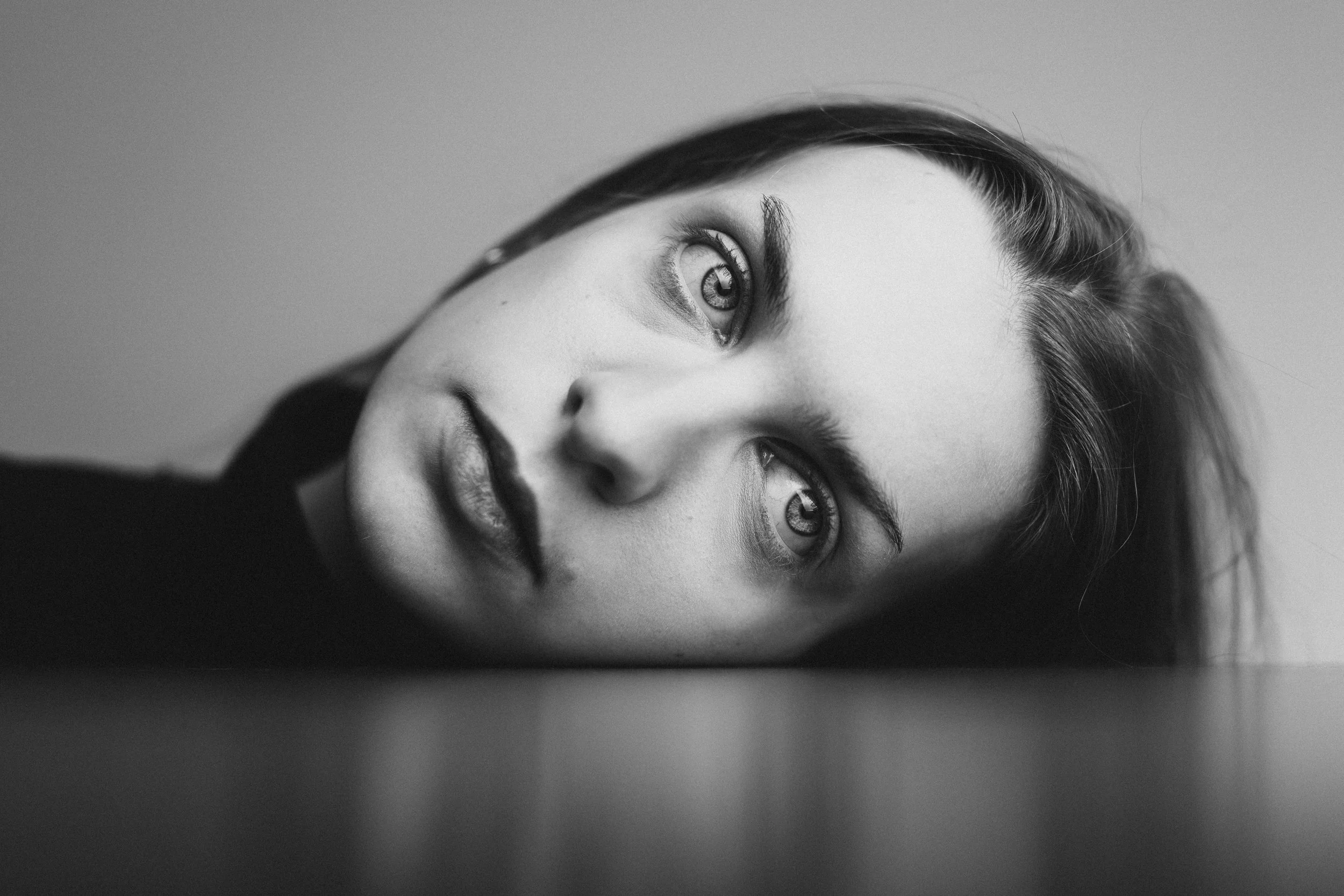 Schwarz-weiß-Fotografie einer Frau, die mit dem Kopf auf einer Oberfläche liegt und direkt in die Kamera blickt.