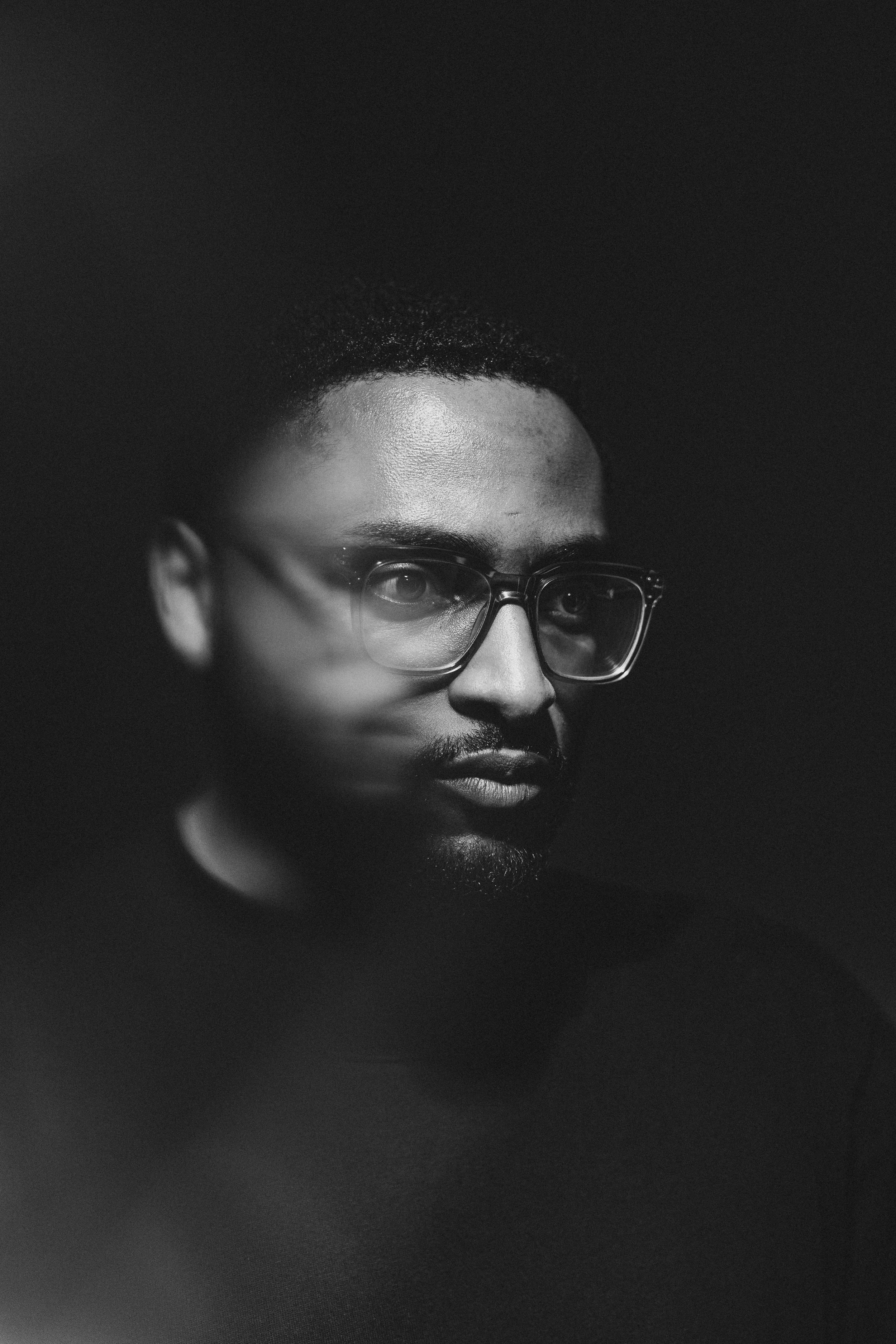 Schwarz-Weiß-Fotografie einer Person mit Brille, Blick nach rechts, dunklem Hintergrund.