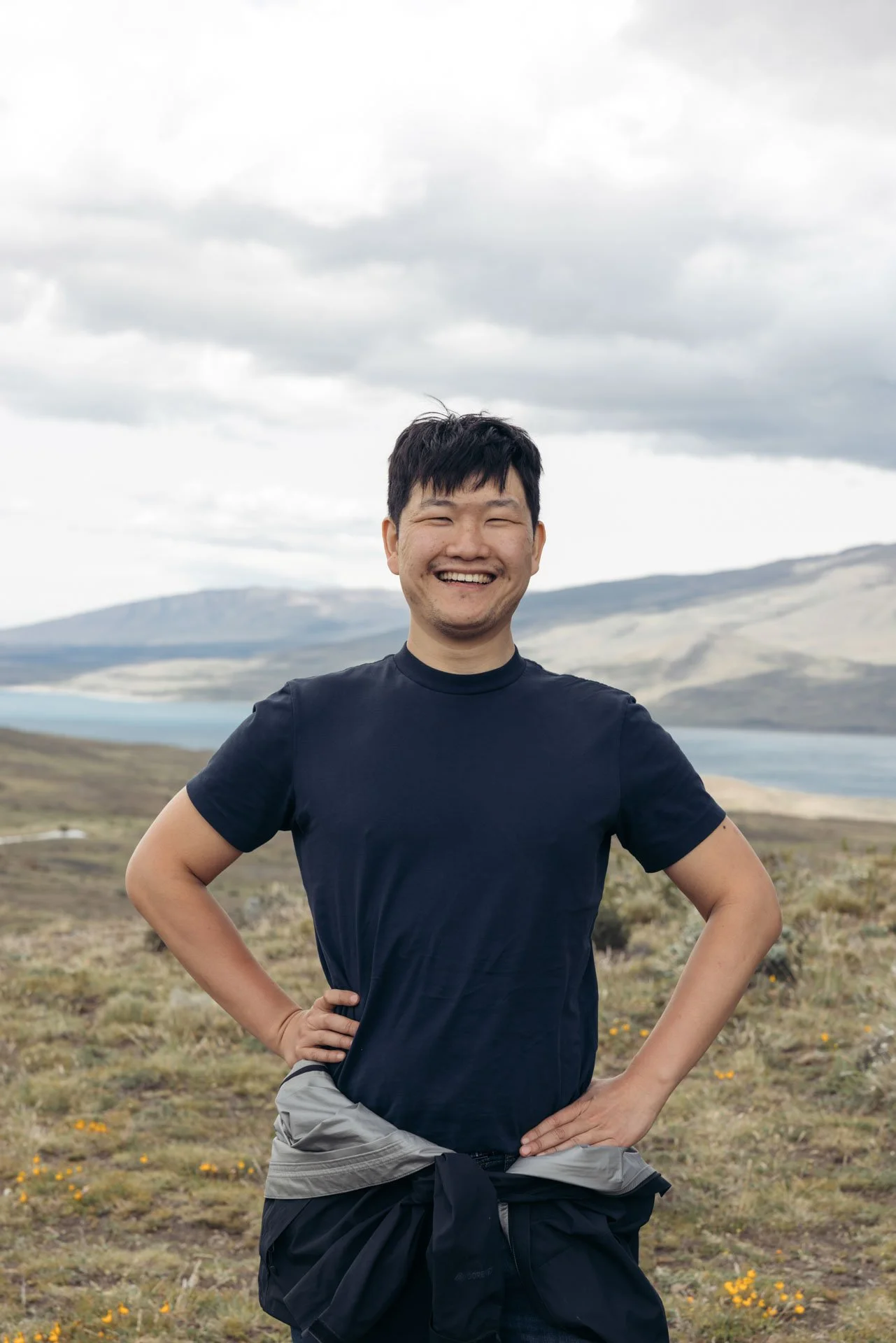 A smiling man standing outdoors in a natural landscape with hills, a lake, and cloudy sky in the background.