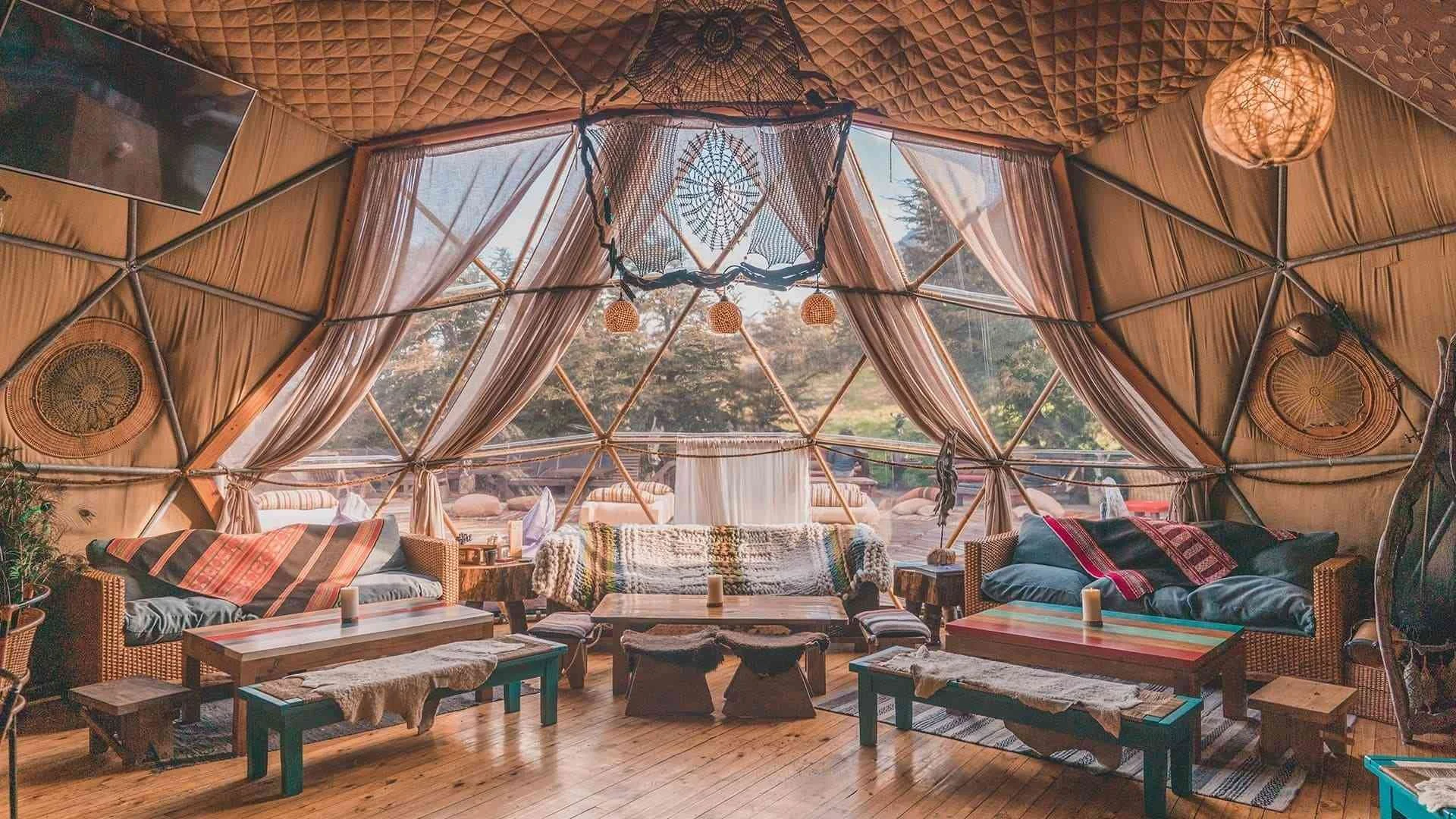 Interior of a geodesic dome with sofas, crochet cushions, wooden tables, and decorative dreamcatchers, overlooking a forested landscape outside.