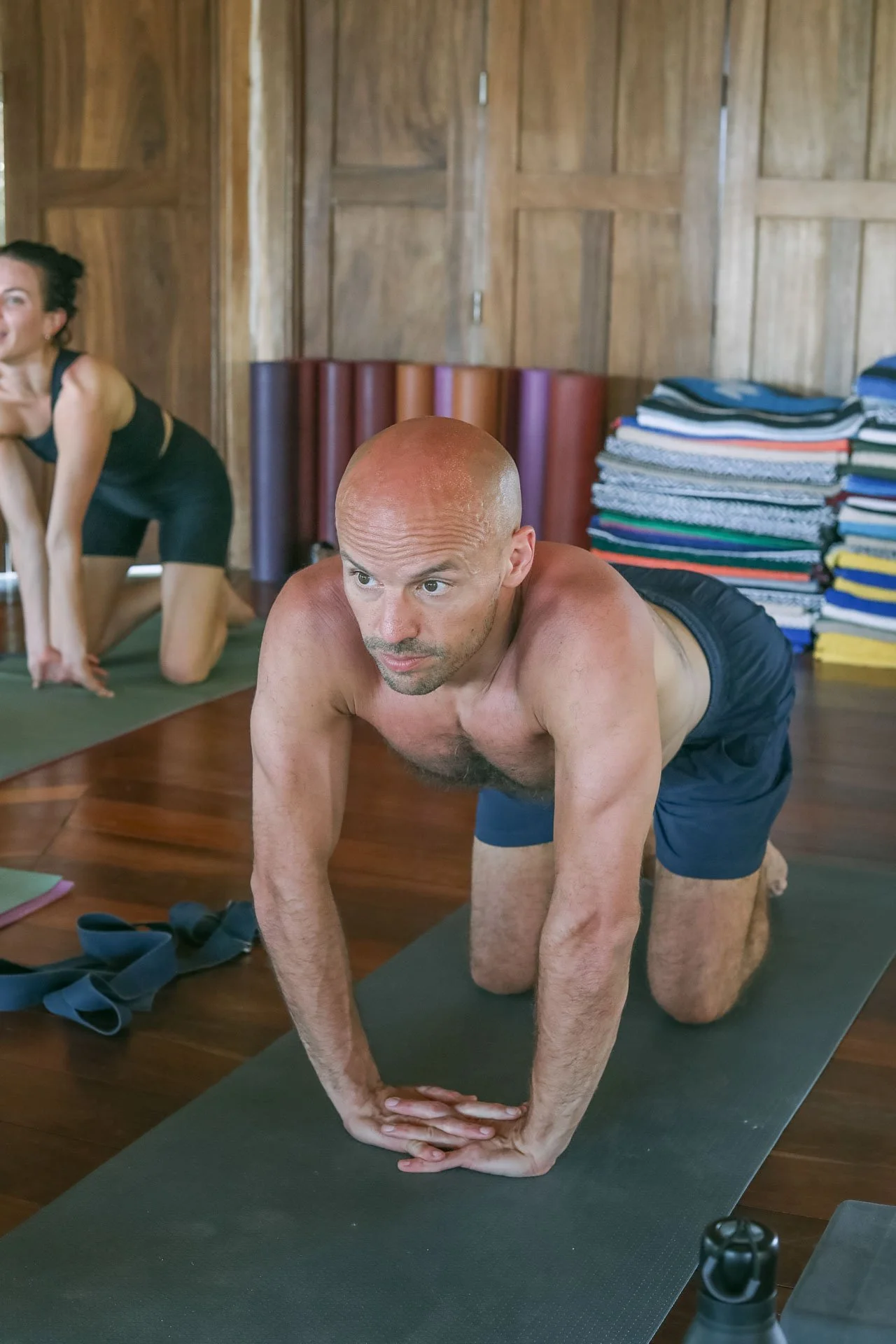 A man with a shaved head and a woman in workout clothes practicing yoga on mats in a wooden room, with the man in a plank position.