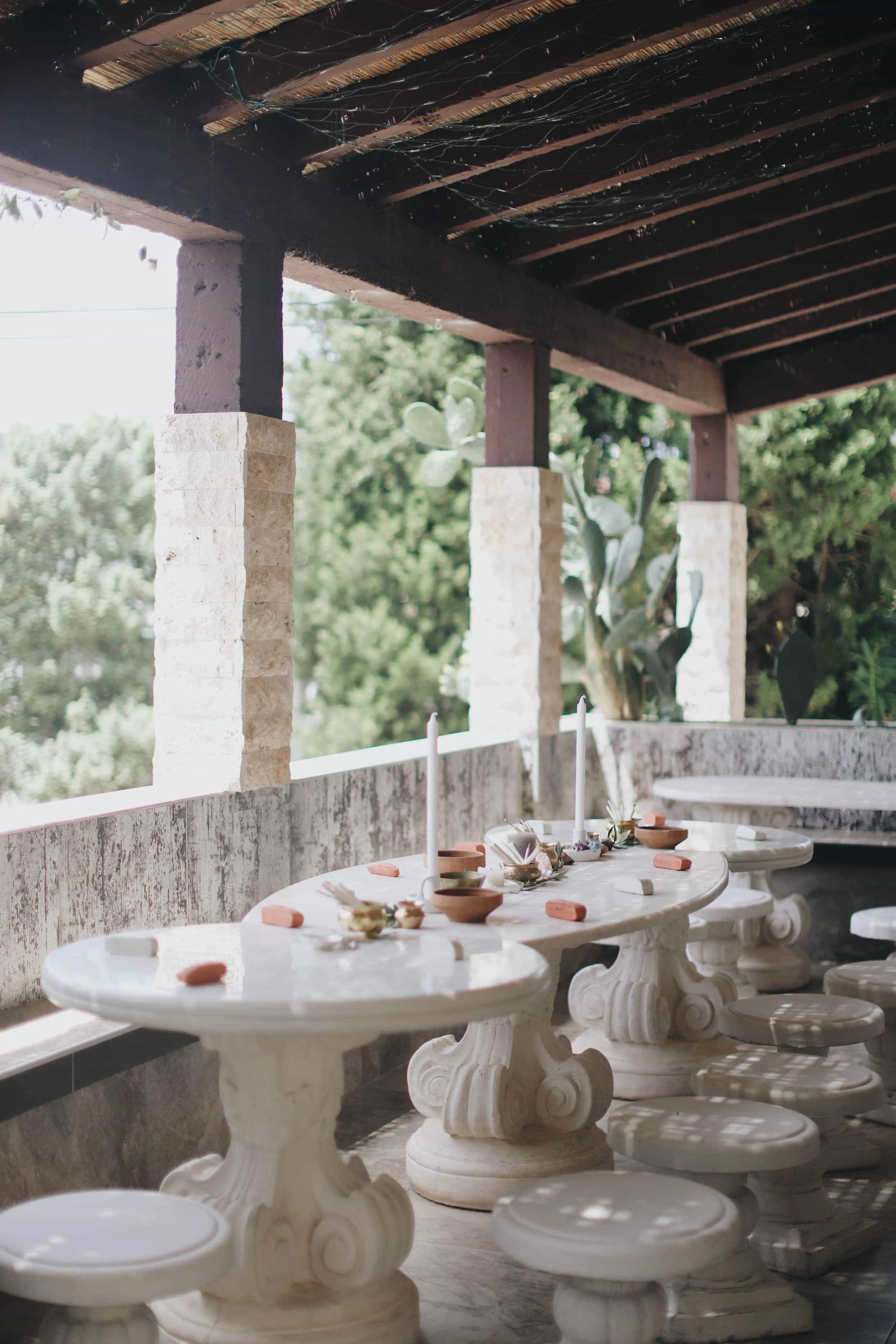 Outdoor patio with three ornate white stone tables, adorned with candles, small bowls, and decorative items, surrounded by matching white stools, under a wooden pergola with hanging vines and greenery in the background.