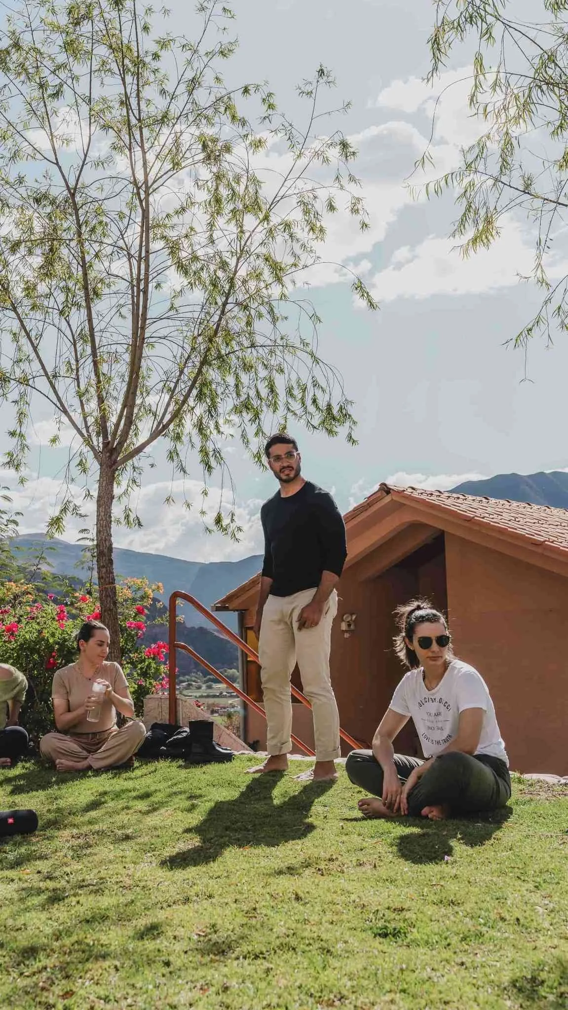 Group of four people relaxing outside on a sunny day with a tree, mountains, and a house in the background. One woman is sitting on the grass, another woman is sitting with a drink, and a man is standing nearby. The fourth person, a woman, is sitting