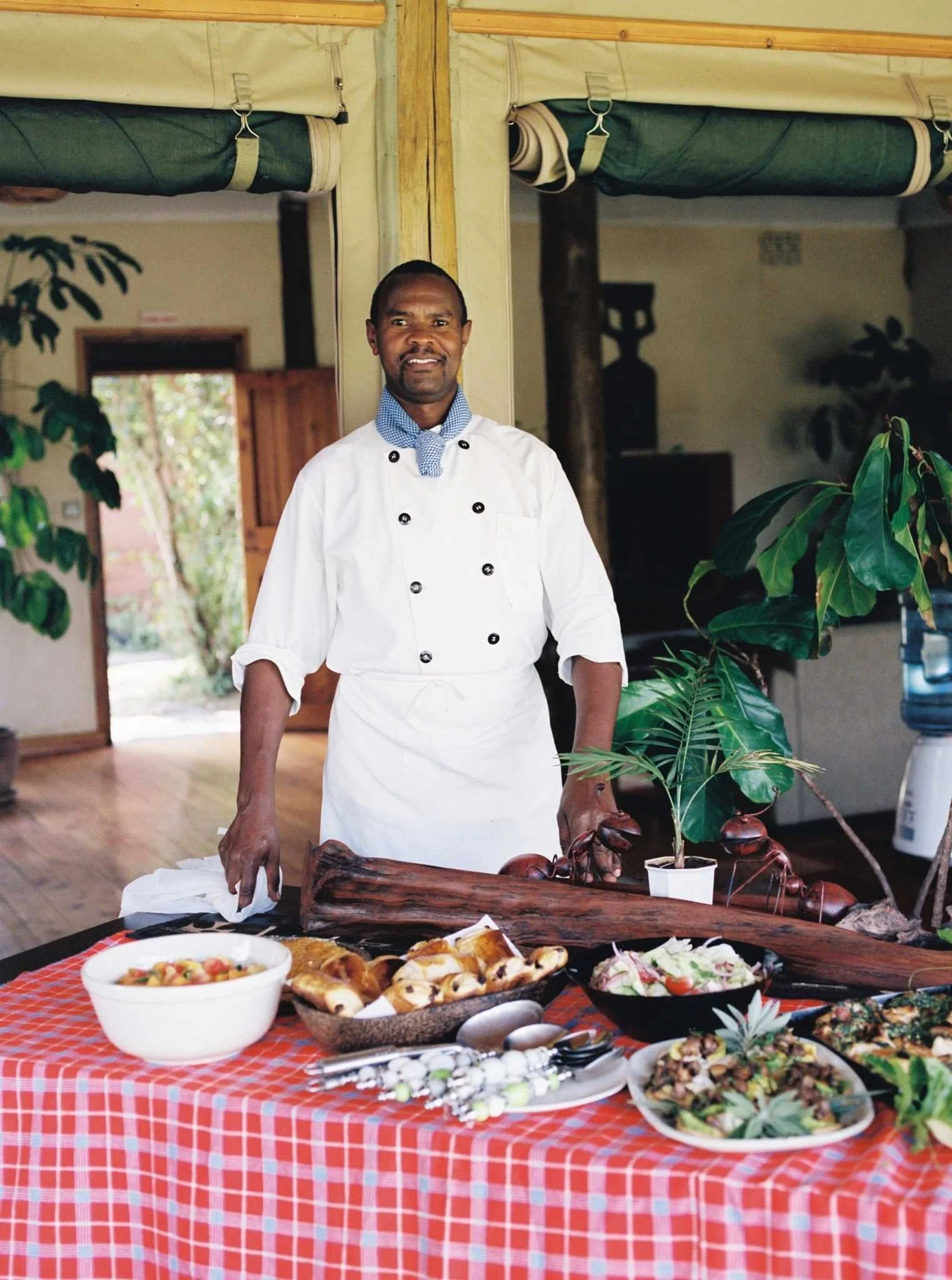 Chef standing behind a table with food, wearing a white chef uniform and a blue checkered neckerchief in a cozy, plant-filled restaurant or kitchen.