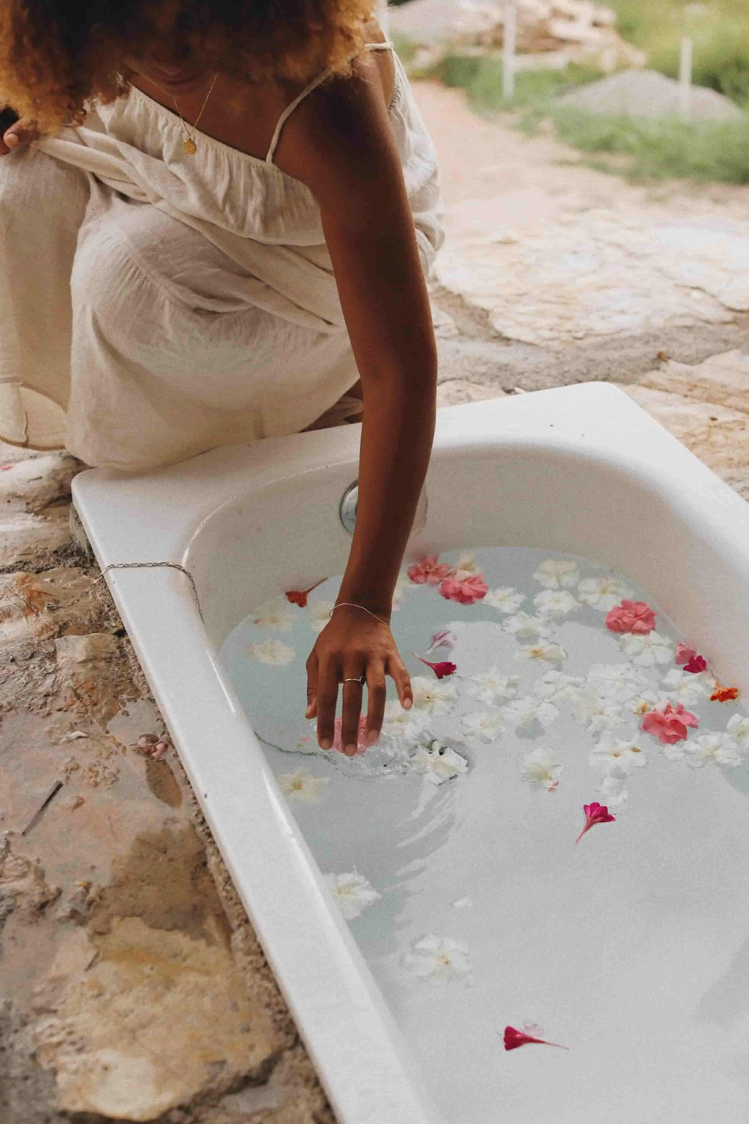 A woman in a beige dress is kneeling by a white basin filled with water and floating flower petals, touching the water with her hand.