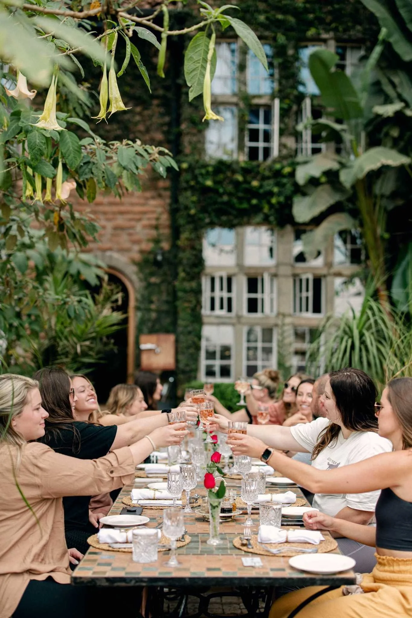 Group of women enjoying a toast at a long outdoor dining table, set with plates, glasses, and flower centerpieces, in a garden setting with lush greenery and a brick building in the background.
