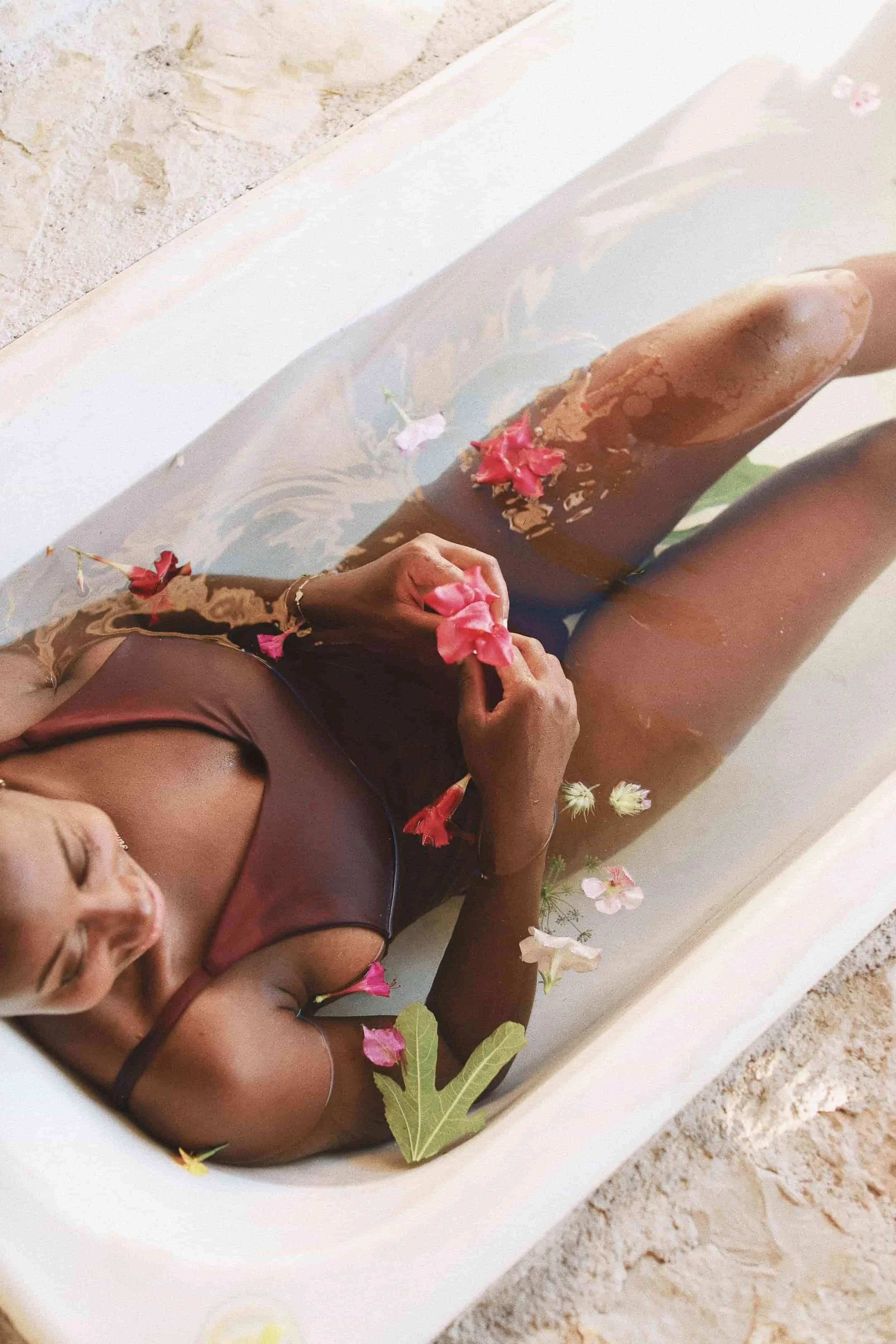 Woman lying in a bathtub filled with water, flowers, and leaves, seen from above.