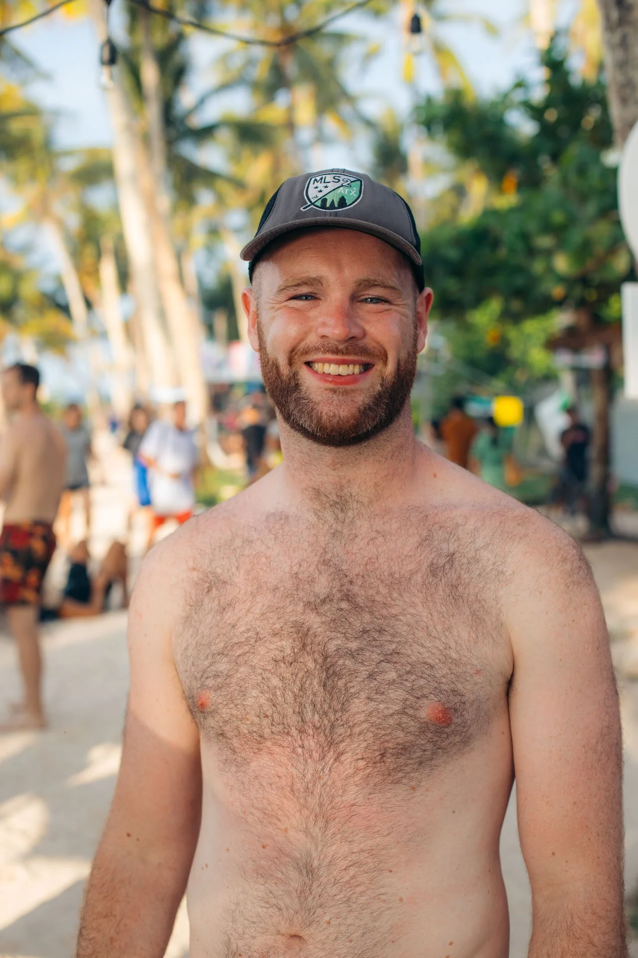 A shirtless man smiling at the camera on a beach with trees and other people in the background.