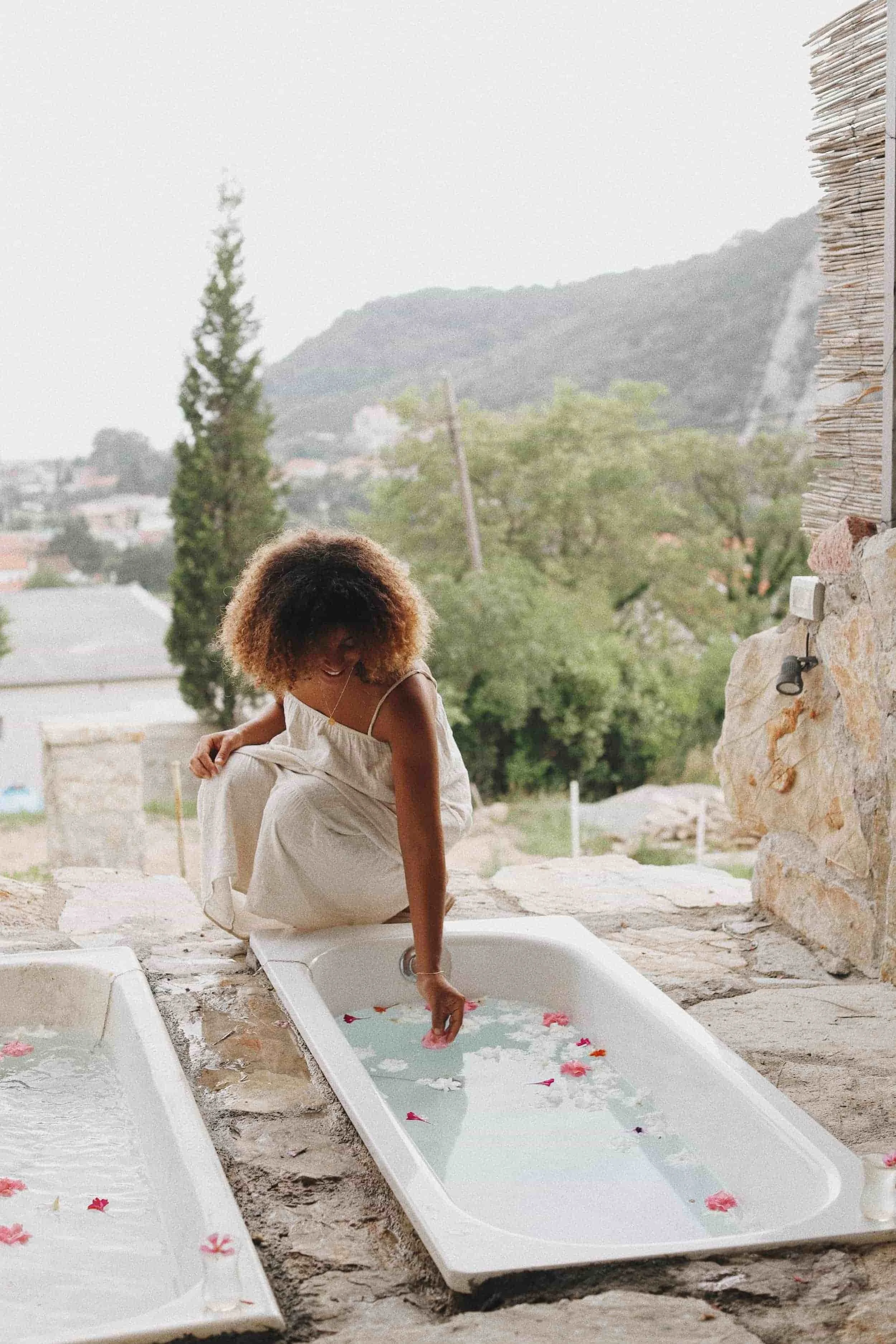 A woman kneeling beside a bathtub, touching the water which is decorated with floating flowers, on a stone patio with a mountainous landscape in the background.