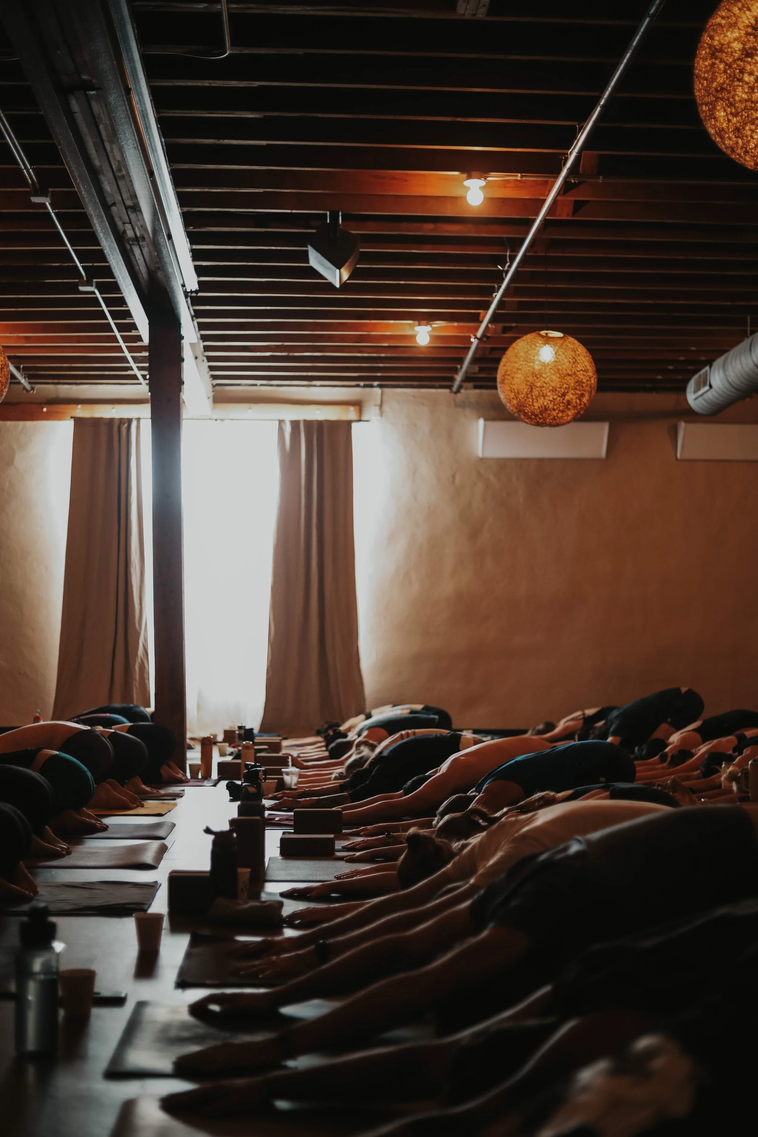 People participating in a yoga class in a dimly lit studio with wooden ceiling beams, soft lighting, and curtains covering a window.