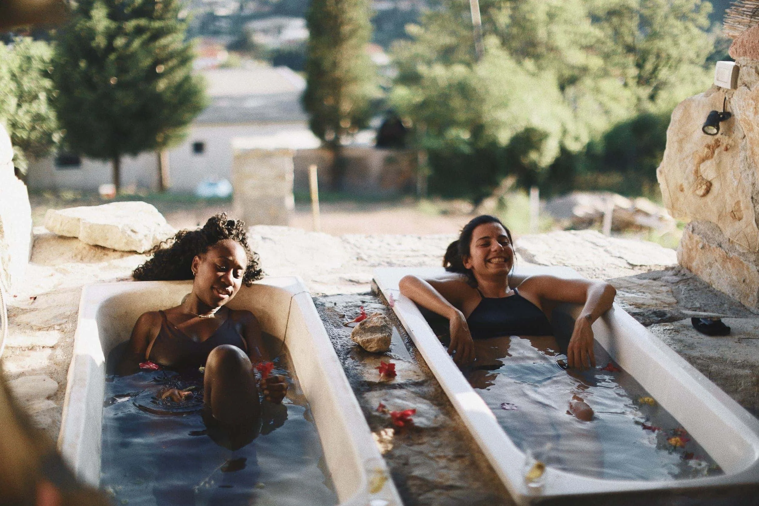 Two women are relaxing in outdoor soaking tubs filled with water and flower petals, surrounded by stone and natural outdoor scenery.