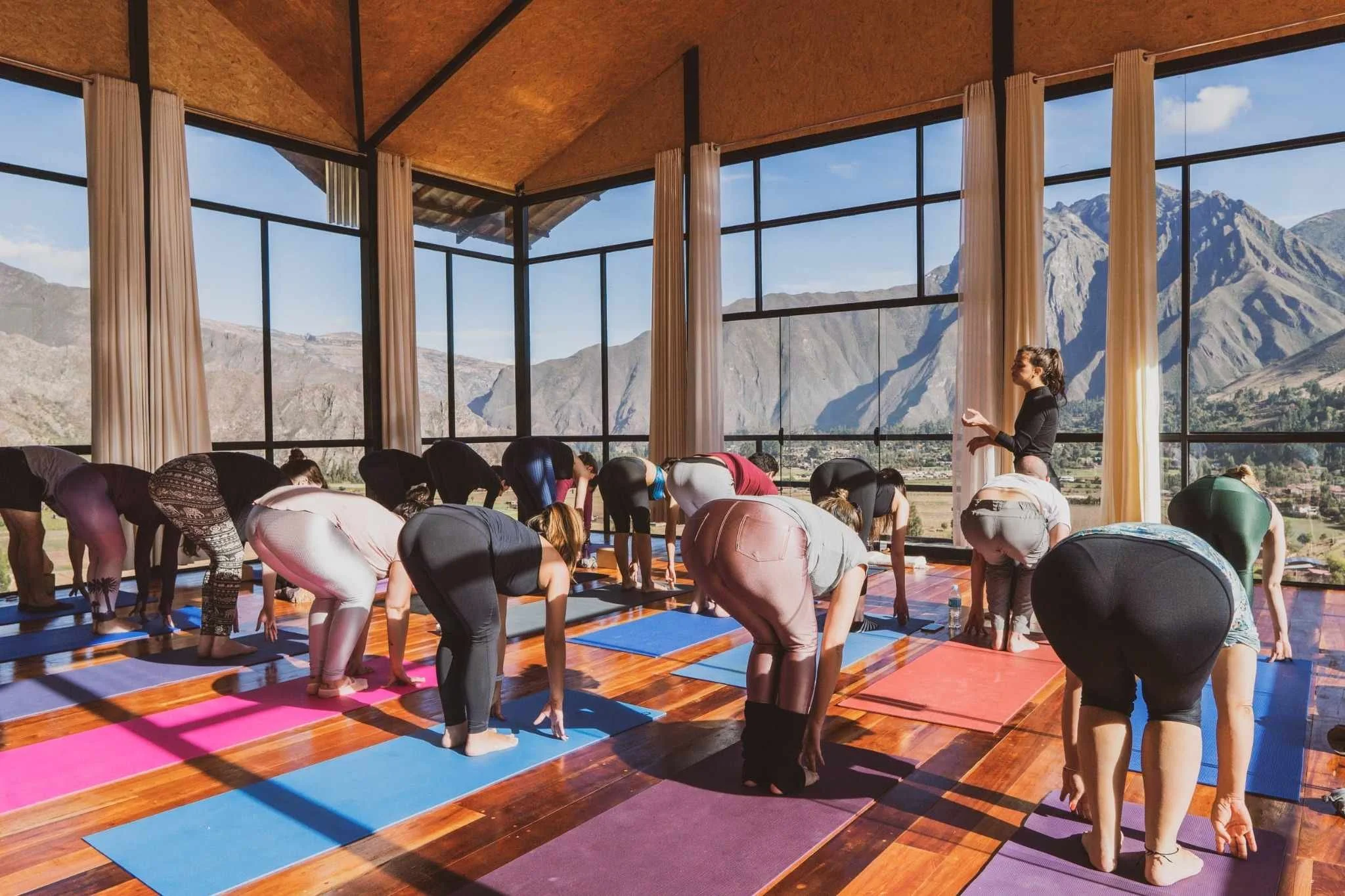 Yoga class in a studio with large windows showing mountain views, participants are bending forward on yoga mats, instructor standing nearby.