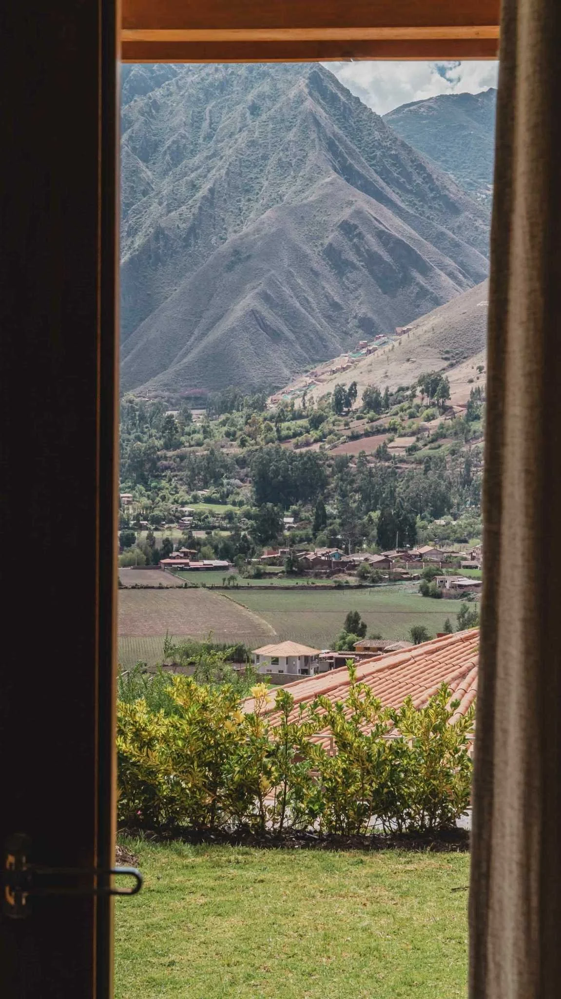 View of a mountainous landscape through an open door with beige curtains, showing green trees, houses, agricultural fields, and large mountains in the background.