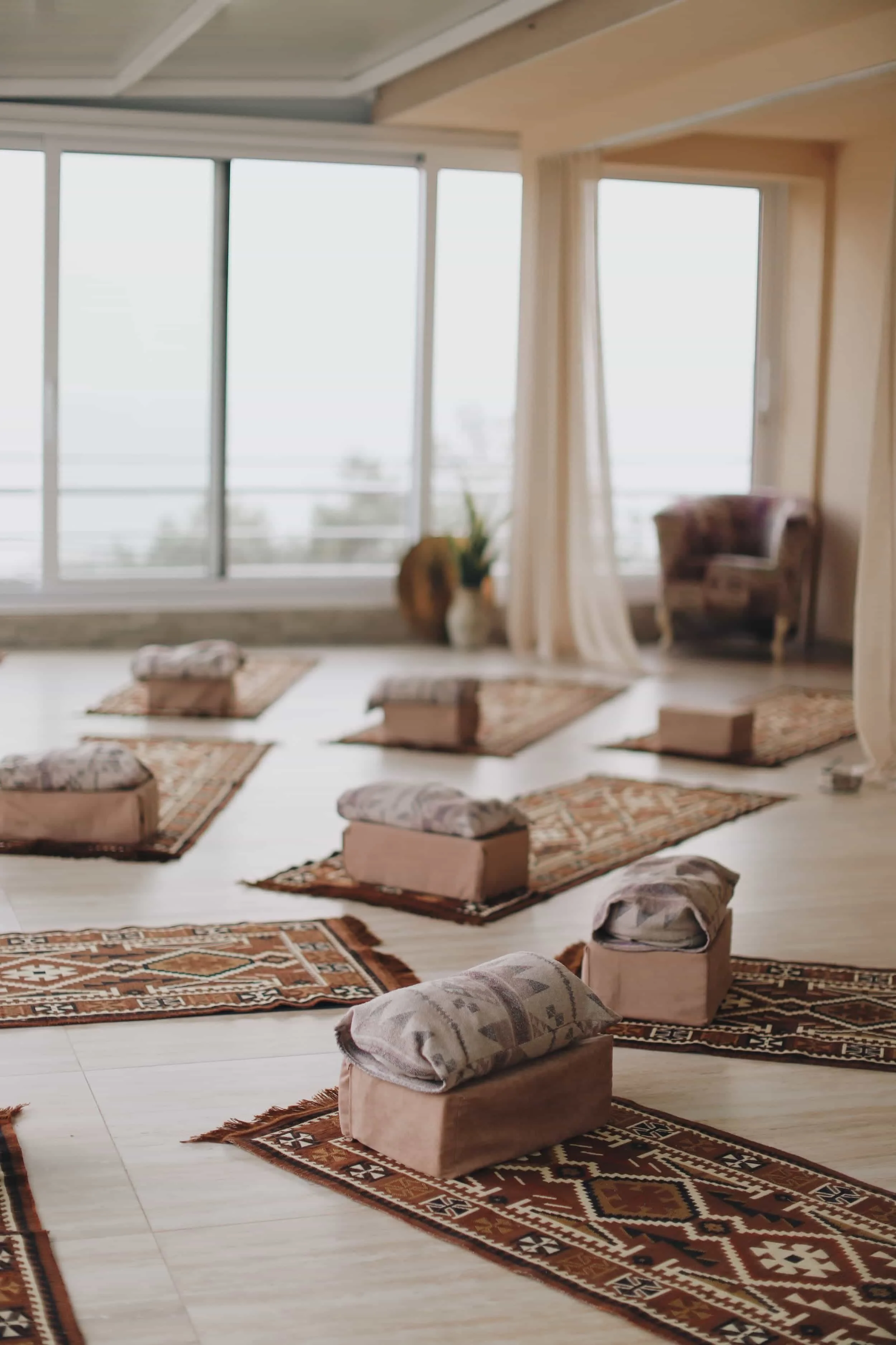 A serene indoor space set up for a yoga or meditation class with cushions and rugs on a wooden floor near large windows that let in natural light.