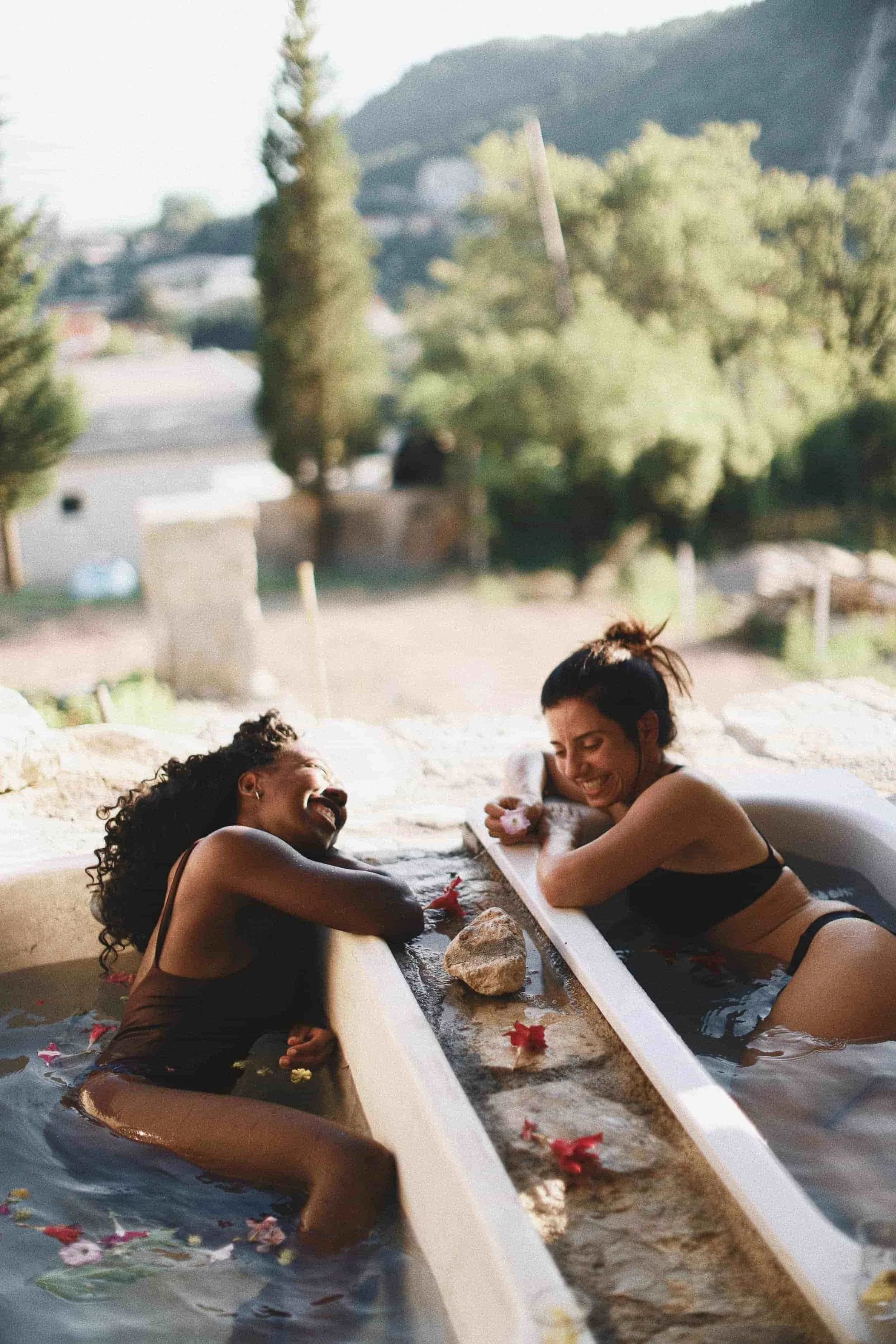 Two women in swimsuits laughing and relaxing in outdoor hot spring bath surrounded by flower petals, with mountains and trees in the background.