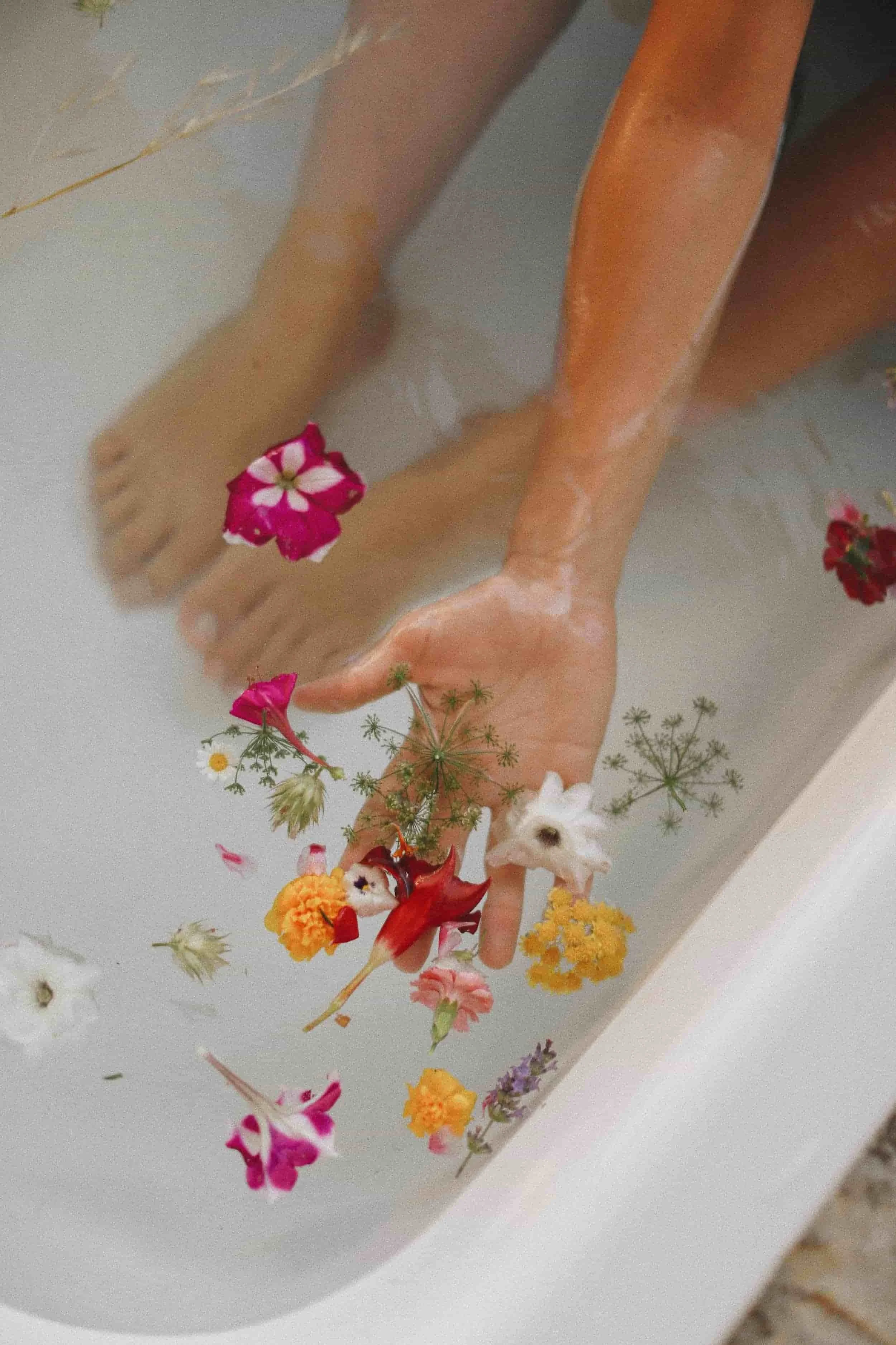 Person submerged in a bathtub filled with water and floating colorful flowers, with one finger touching the water's surface.