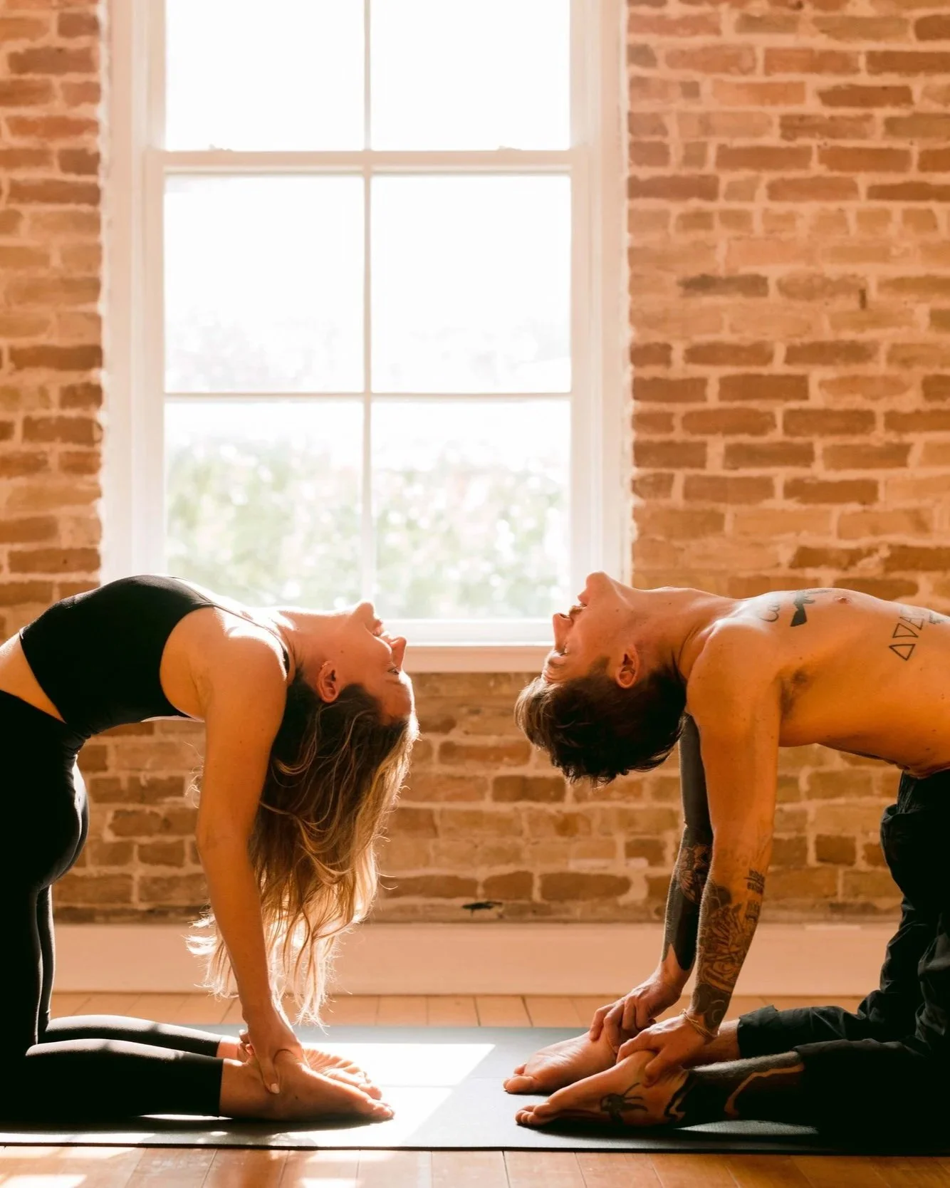 Two people practicing yoga in a room with brick walls, facing each other in a bow pose, with their foreheads touching and holding their feet.