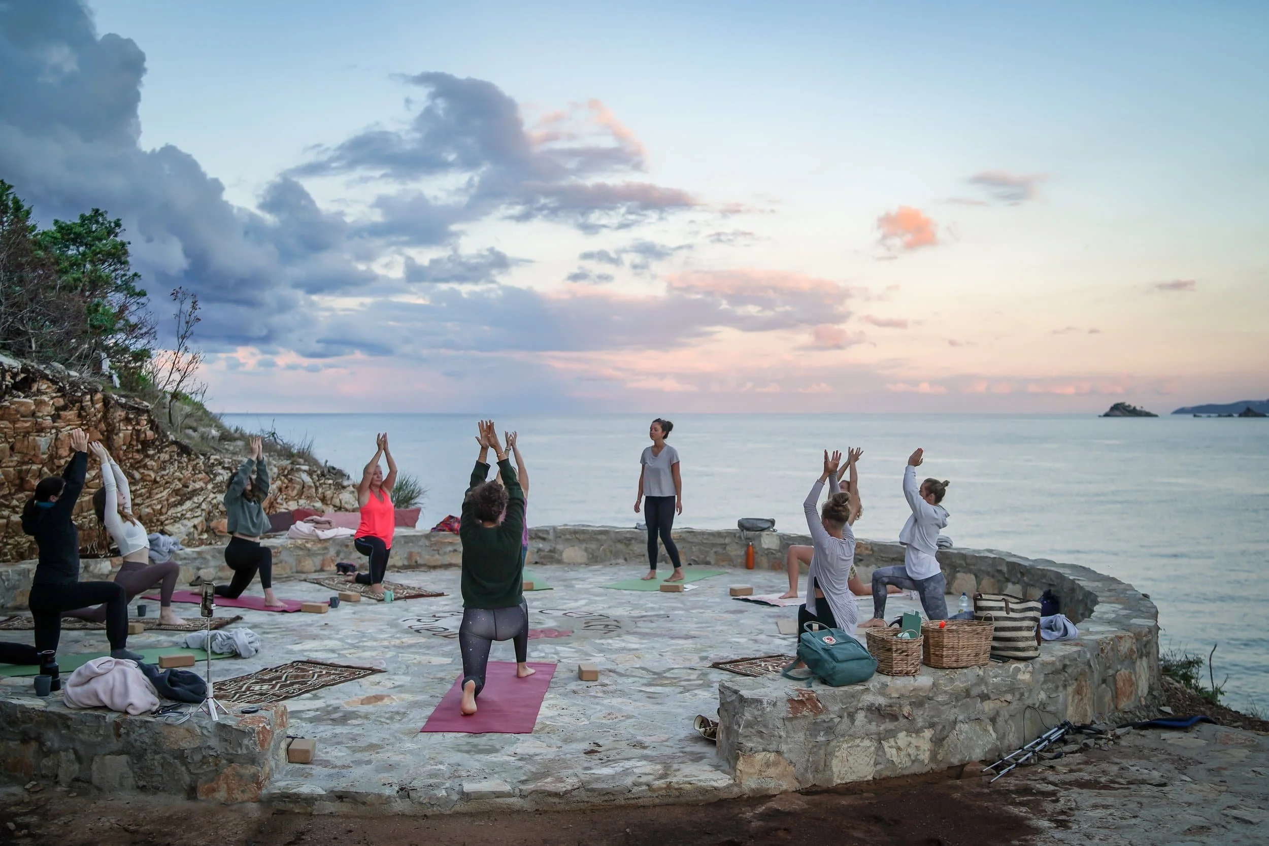 Group of people practicing yoga outdoors on a stone platform by the water at sunset.