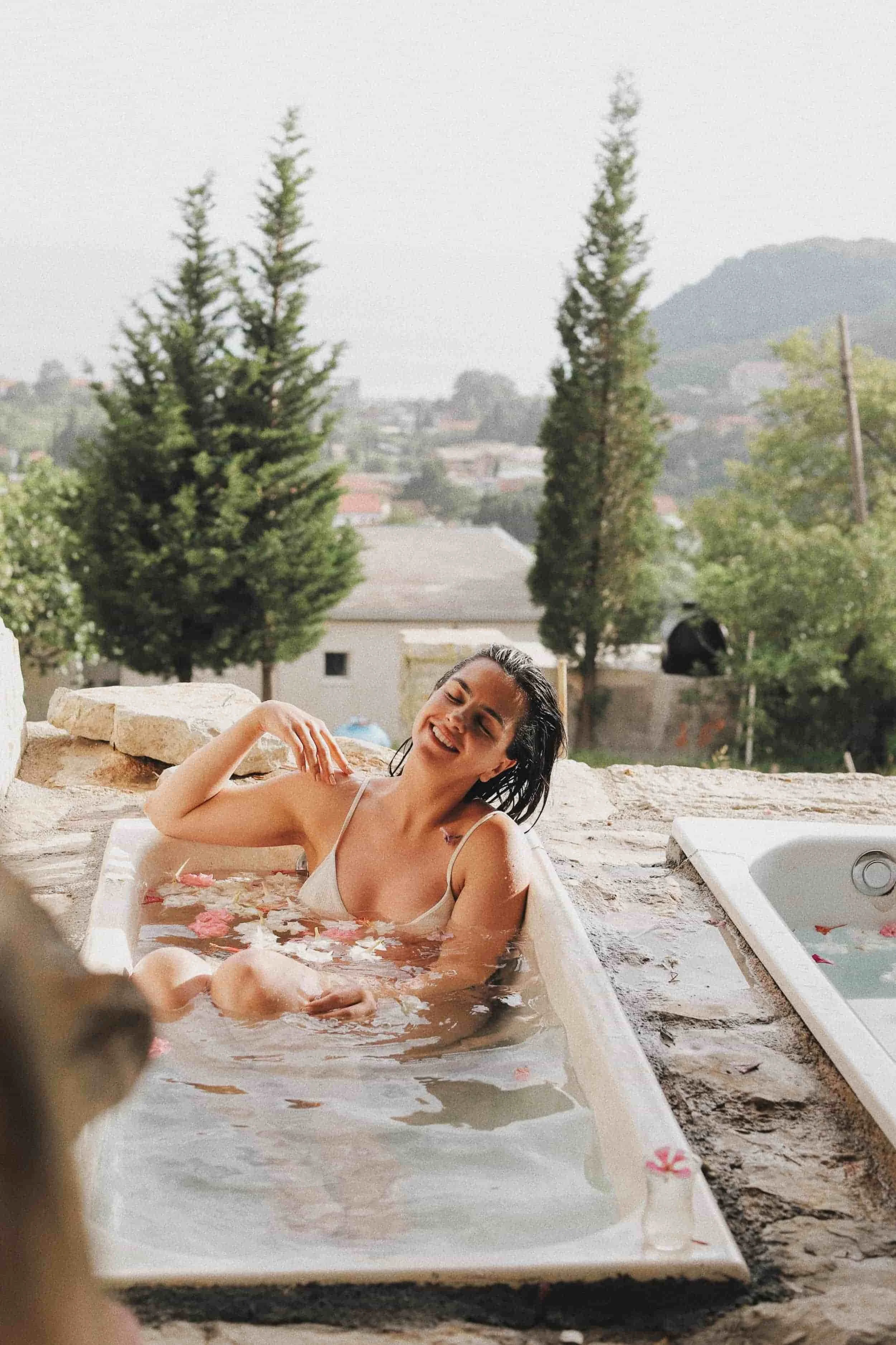 A woman relaxing in a bathtub outdoors on a stone patio with trees and hills in the background, smiling and enjoying a bath with flower petals.
