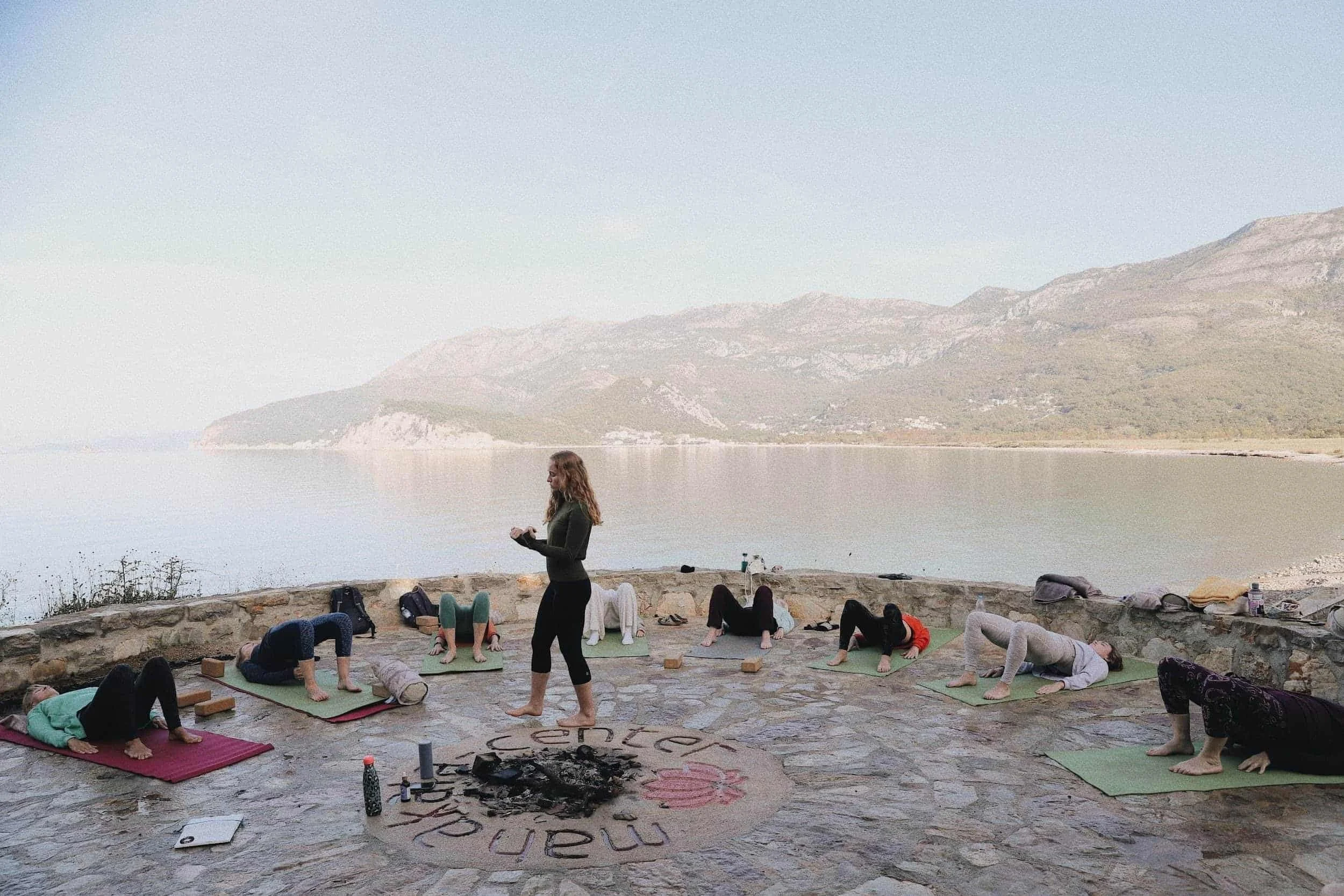 Group of people practicing yoga outdoors on a stone patio near a body of water with mountains in the background, led by an instructor standing in the center.