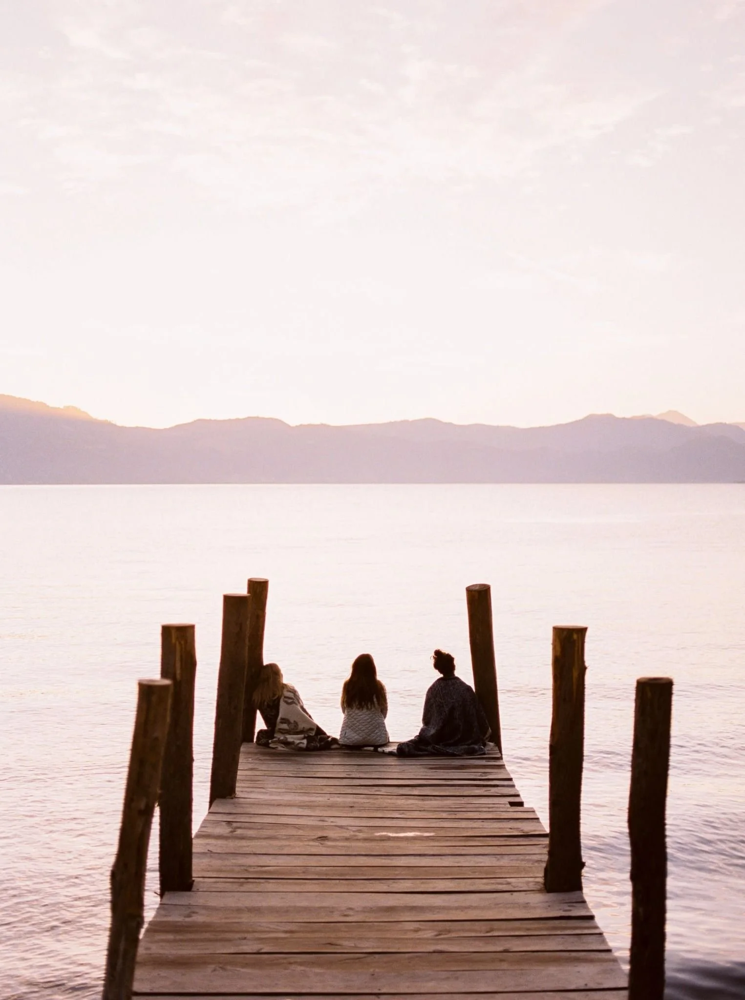 Three people sitting on a wooden dock over a calm lake during sunset, with distant mountains and a pastel-colored sky in the background.