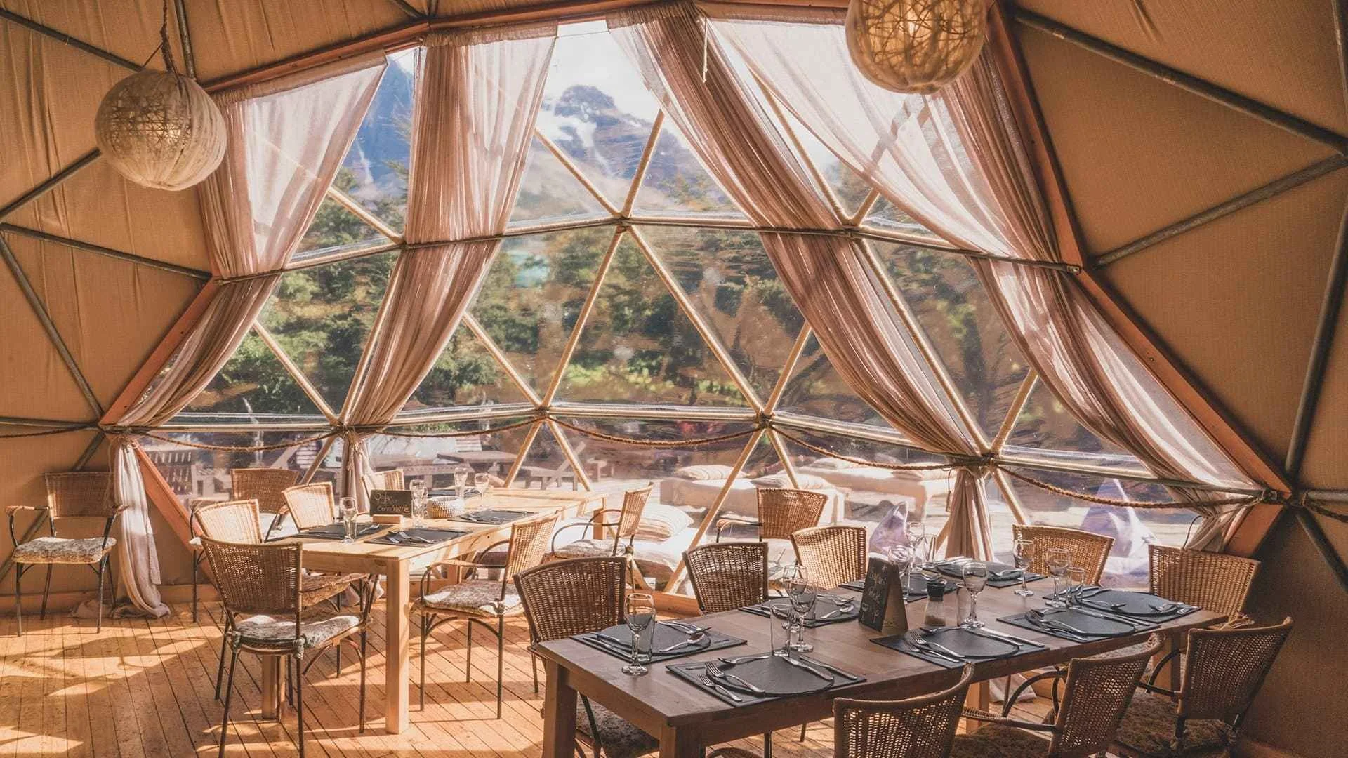 Interior of a glamping tent with large geometric windows, decorated with curtains and hanging paper lanterns, set up for dining with tables, chairs, and tableware.