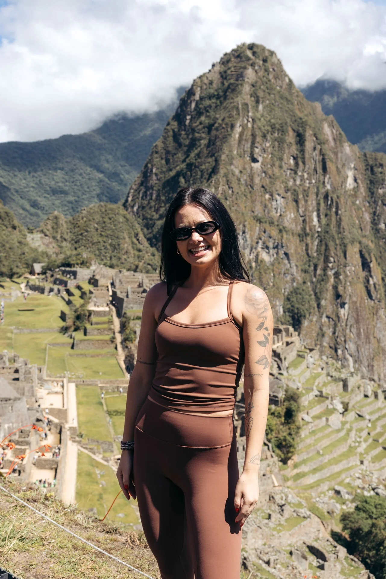 A woman with black hair, sunglasses, and a tan outfit standing in front of Machu Picchu with the mountain and ruins in the background.